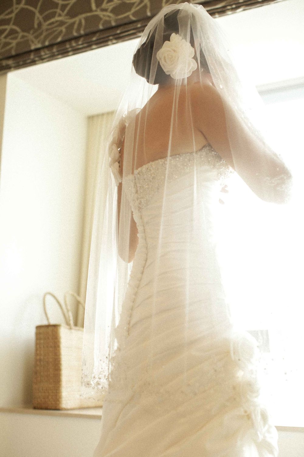 A bride in a white wedding gown and veil stands in soft window light creating an ethereal bridal portrait.