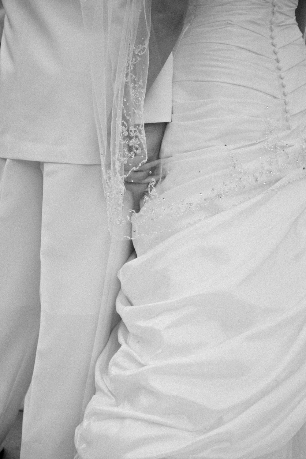 Black and white closeup of a bride and groom holding hands, showing her white wedding dress and his light suit.