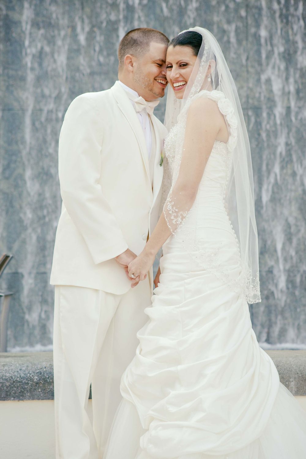 A bride in white wedding dress and veil shares a tender moment with groom in white suit against textured gray wall.