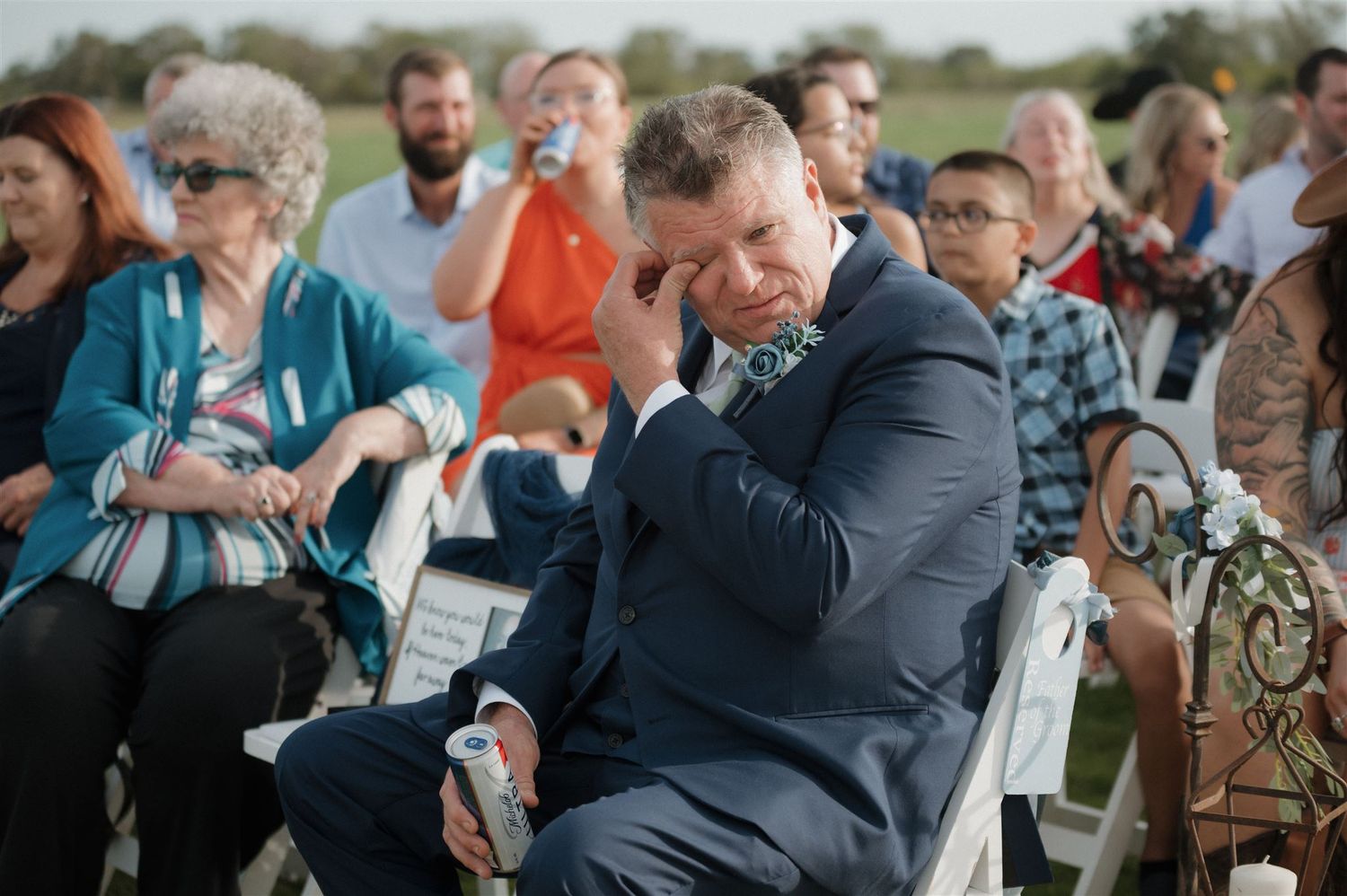 Guests seated in chairs at an outdoor ceremony watching someone in a navy suit showing emotion.
