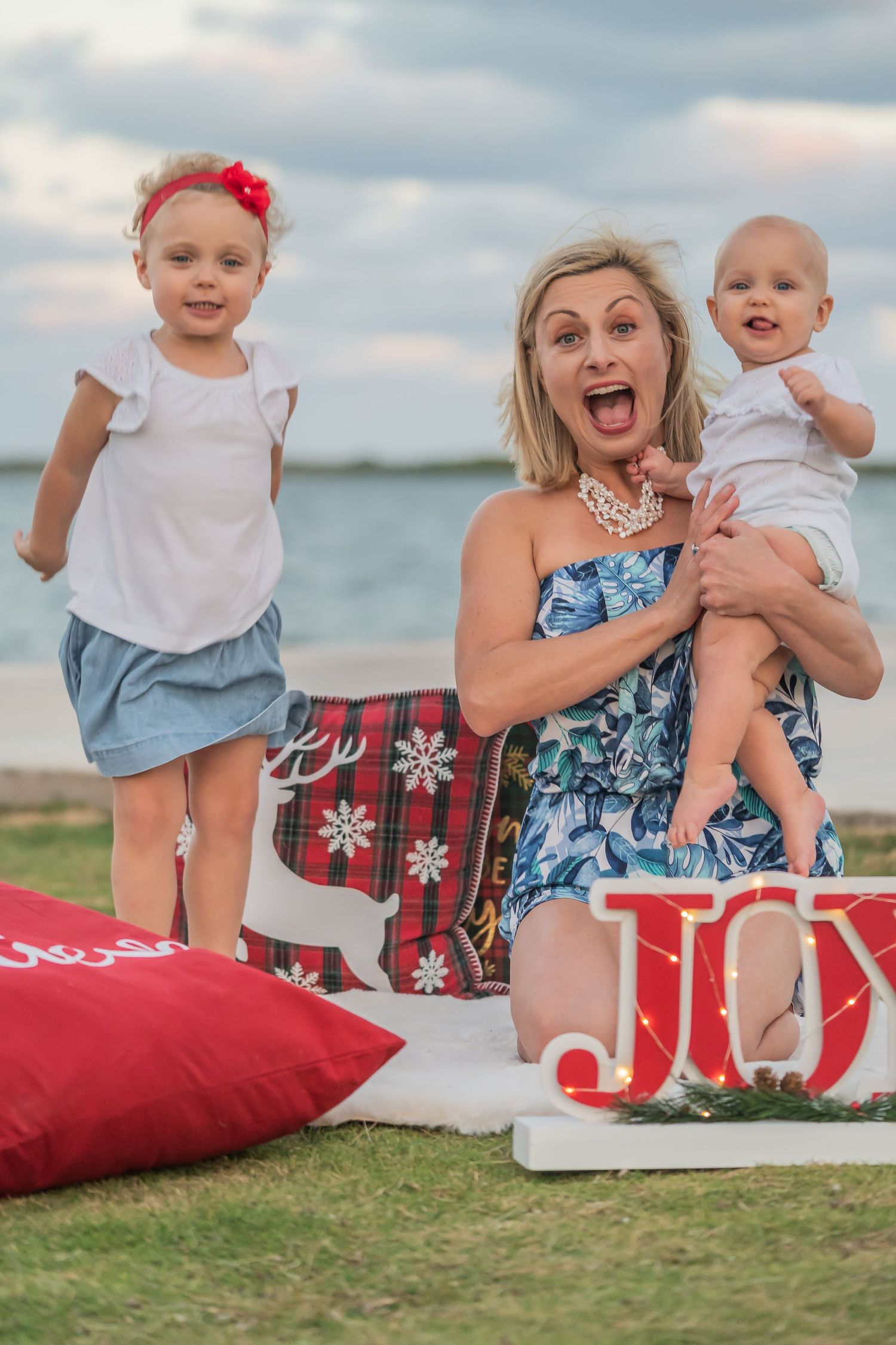 Holiday photo session on grass with JOY letters and festive red pillows near the ocean.