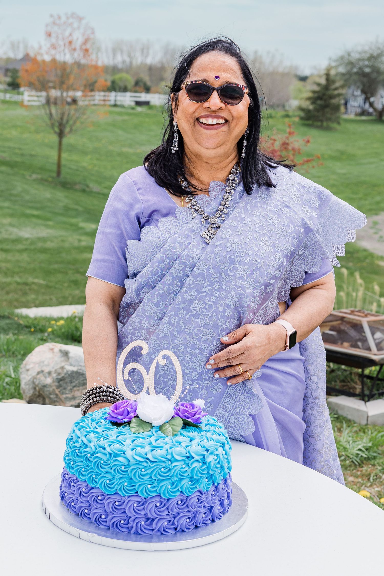 A person in a purple sari stands next to a blue ombre birthday cake decorated with the number 60.