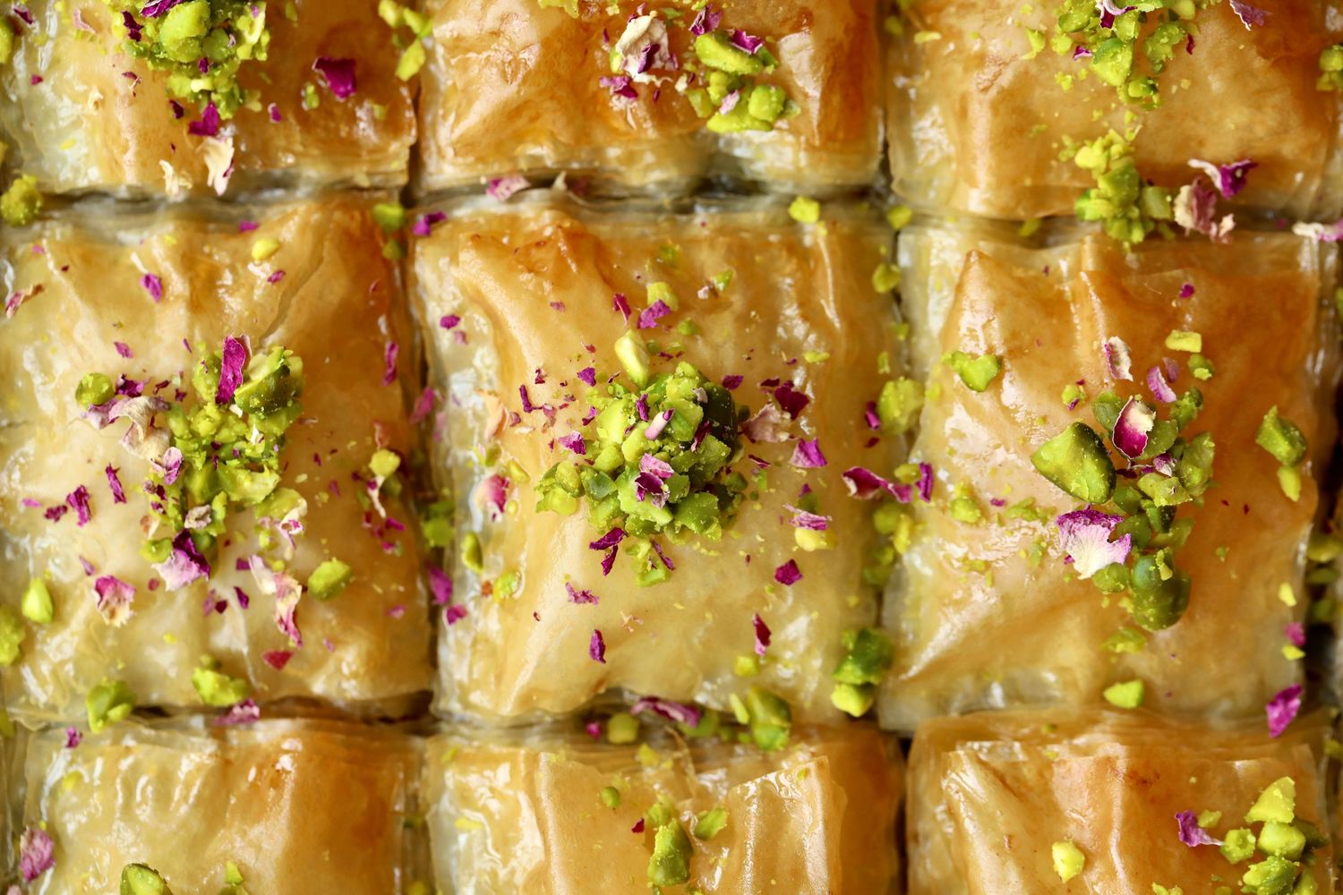 Golden flaky baklava pastry garnished with crushed pistachios and rose petals in a baking pan.