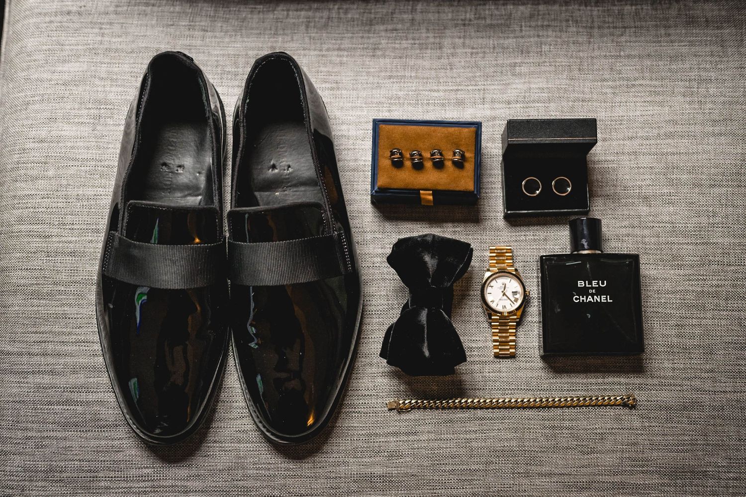 Flat lay of formal men's accessories including patent leather shoes, cufflinks and watch on gray fabric.