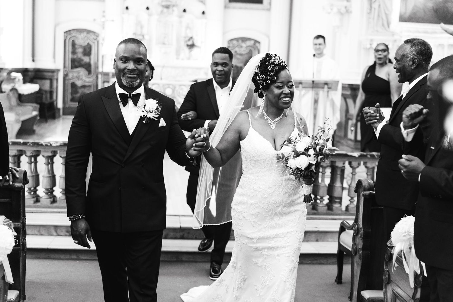 Black and white photo of joyful newlyweds walking down church aisle, bride raising bouquet in celebration after ceremony.