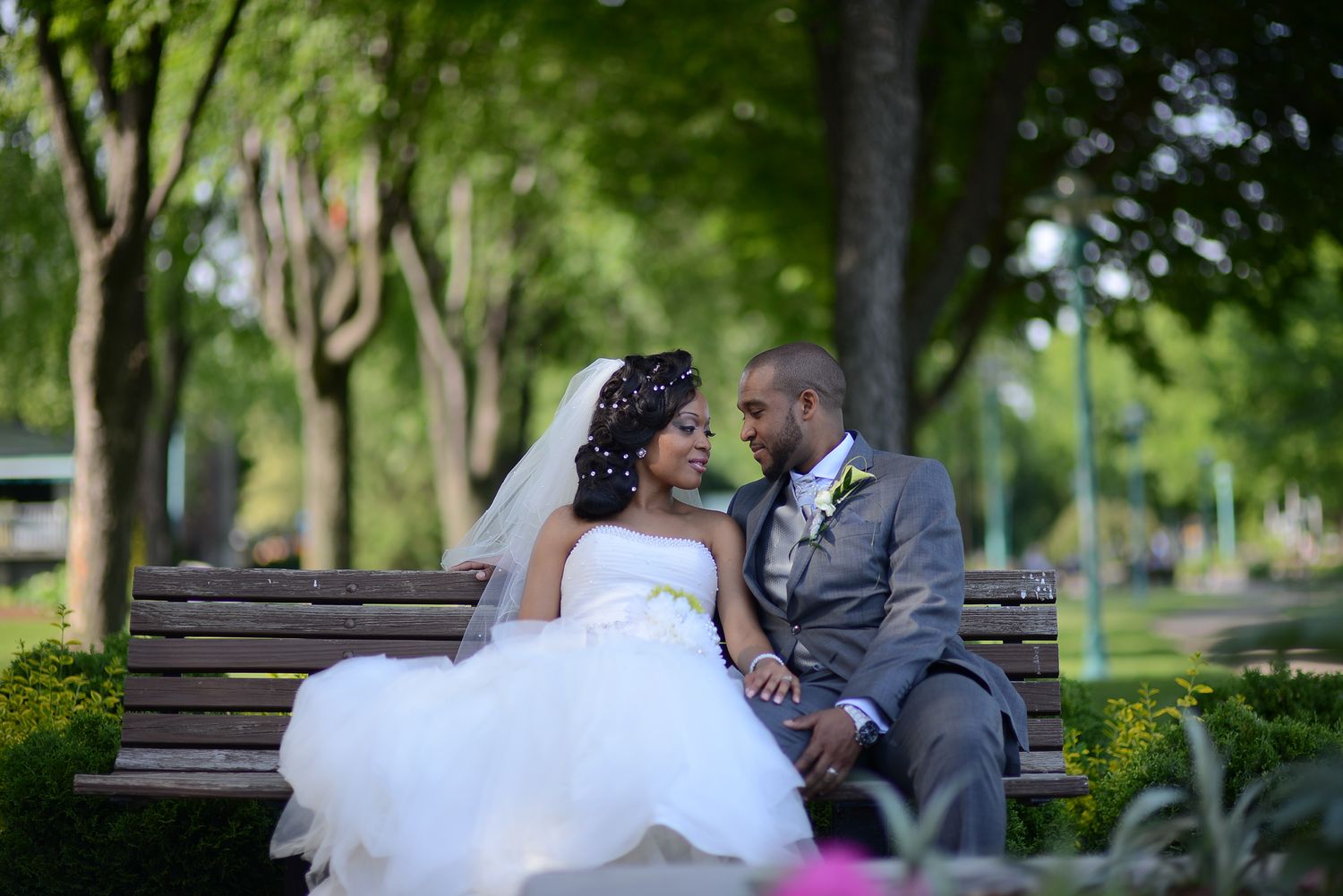 A bride in a white tulle gown and groom in gray suit sit facing each other on a park bench.