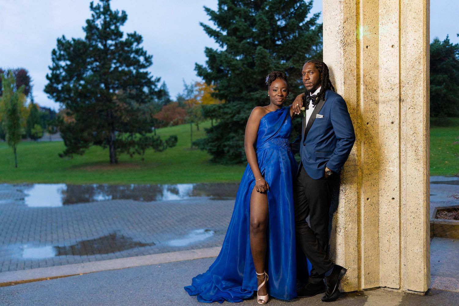 Elegant couple in matching royal blue formal wear posing outdoors near stone columns for prom or gala.