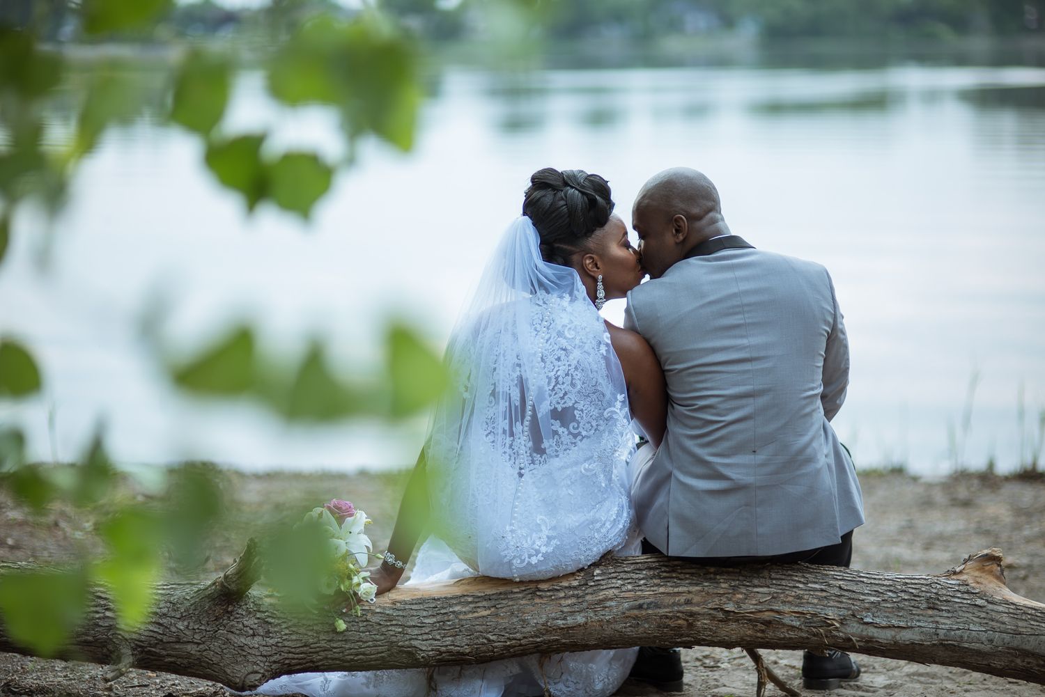 A bride and groom share a kiss while seated on a log near a serene lakeside setting.