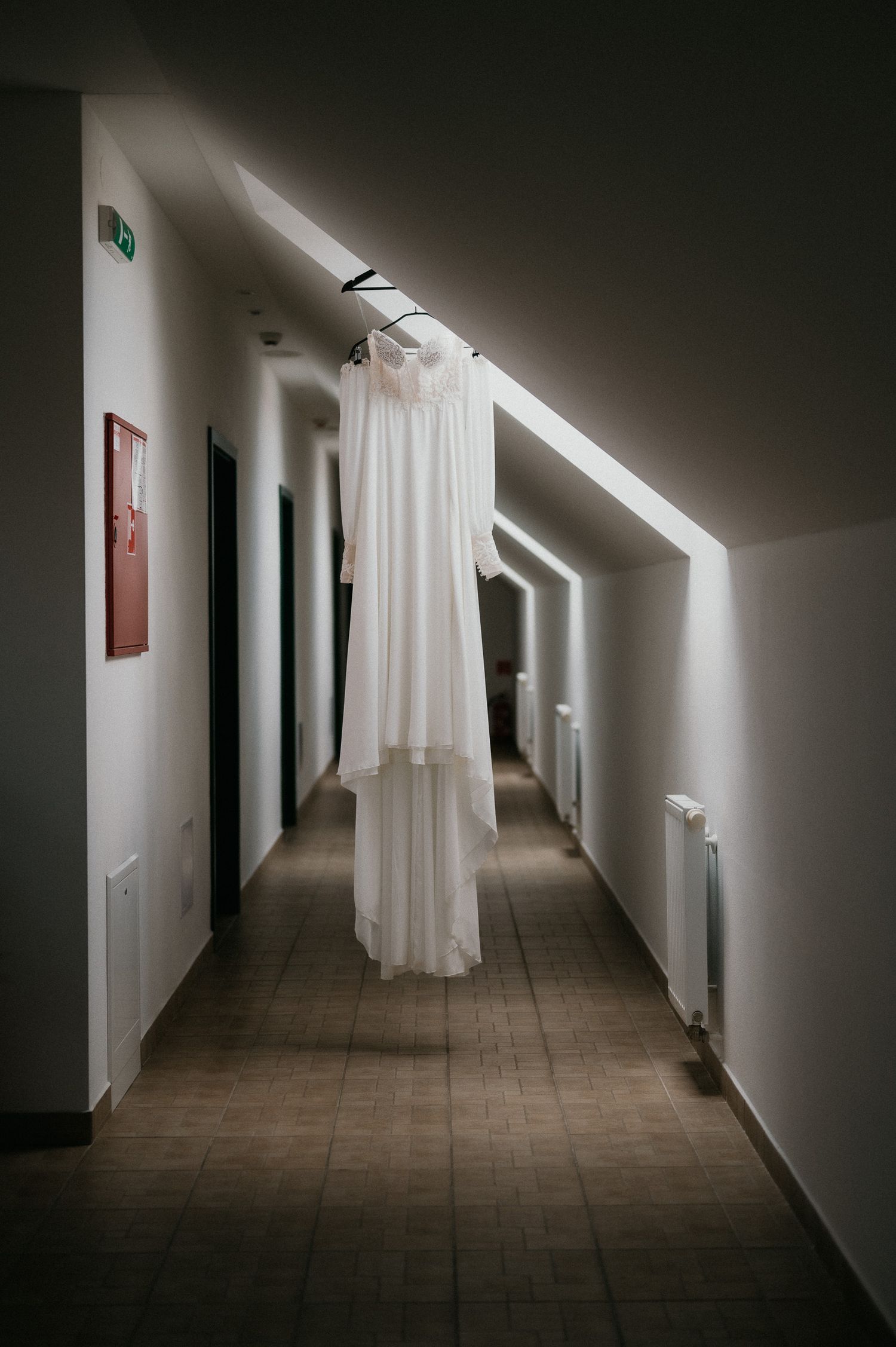 A white wedding dress hangs elegantly in a minimalist hallway with dramatic lighting.