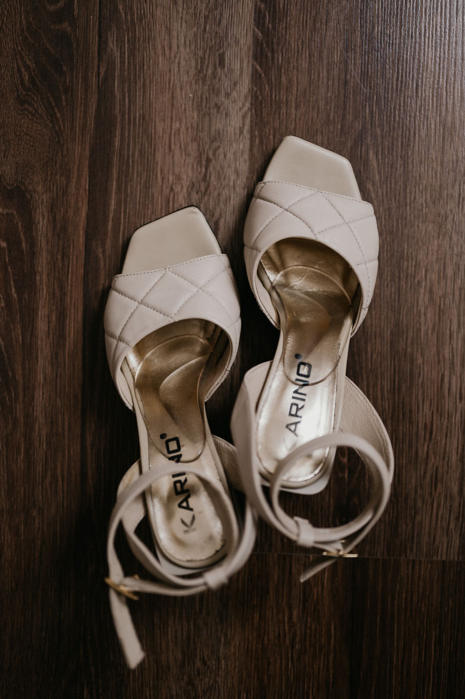 Cream-colored quilted wedding sandals with ankle straps on dark wooden flooring.