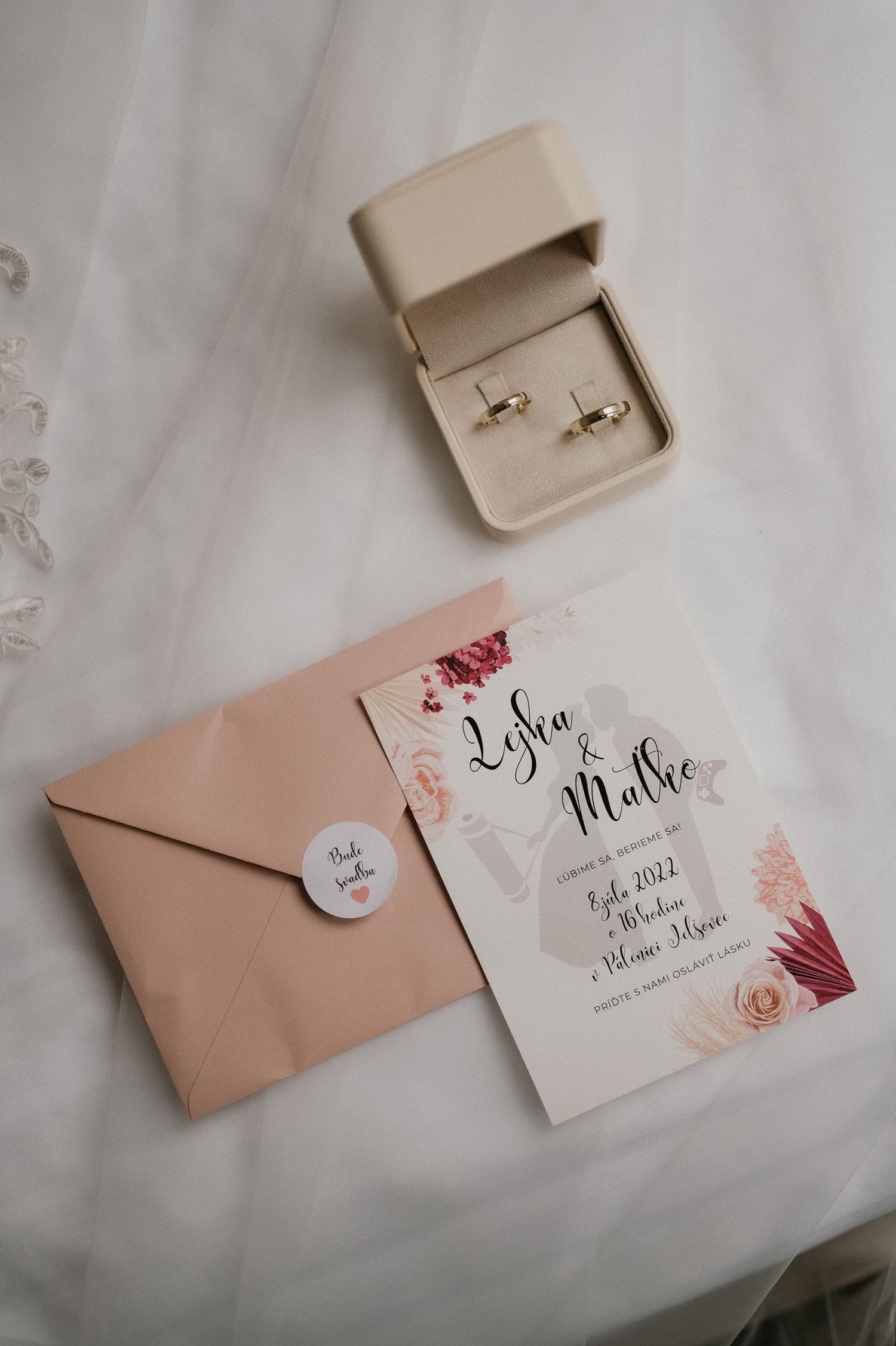 Wedding invitation with floral design and peach envelope beside ring box on white surface.