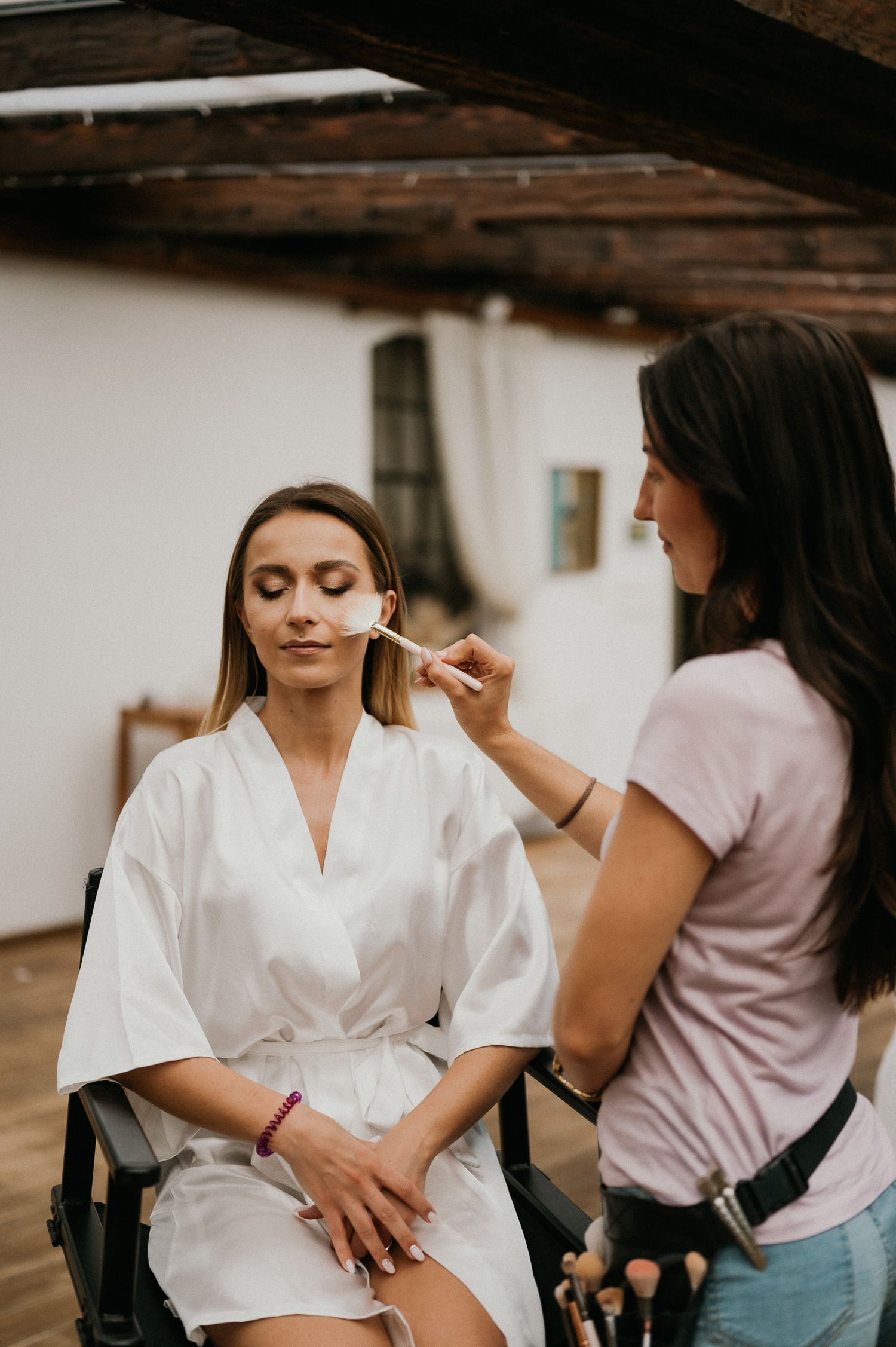 Makeup artist applying cosmetics to a client wearing a white silk robe during wedding preparation.