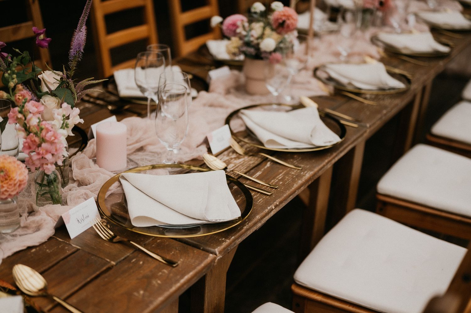 Rustic wooden table set for wedding reception with white plates and floral centerpieces.