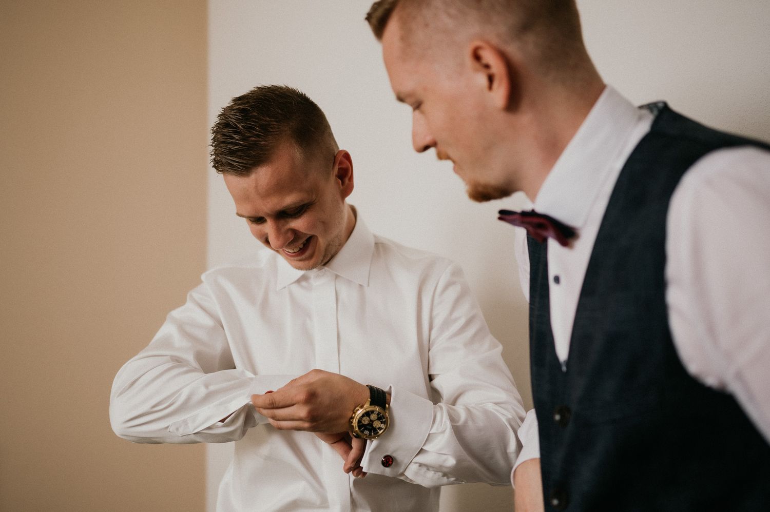 Two groomsmen in white shirts and vests helping each other prepare for the wedding.