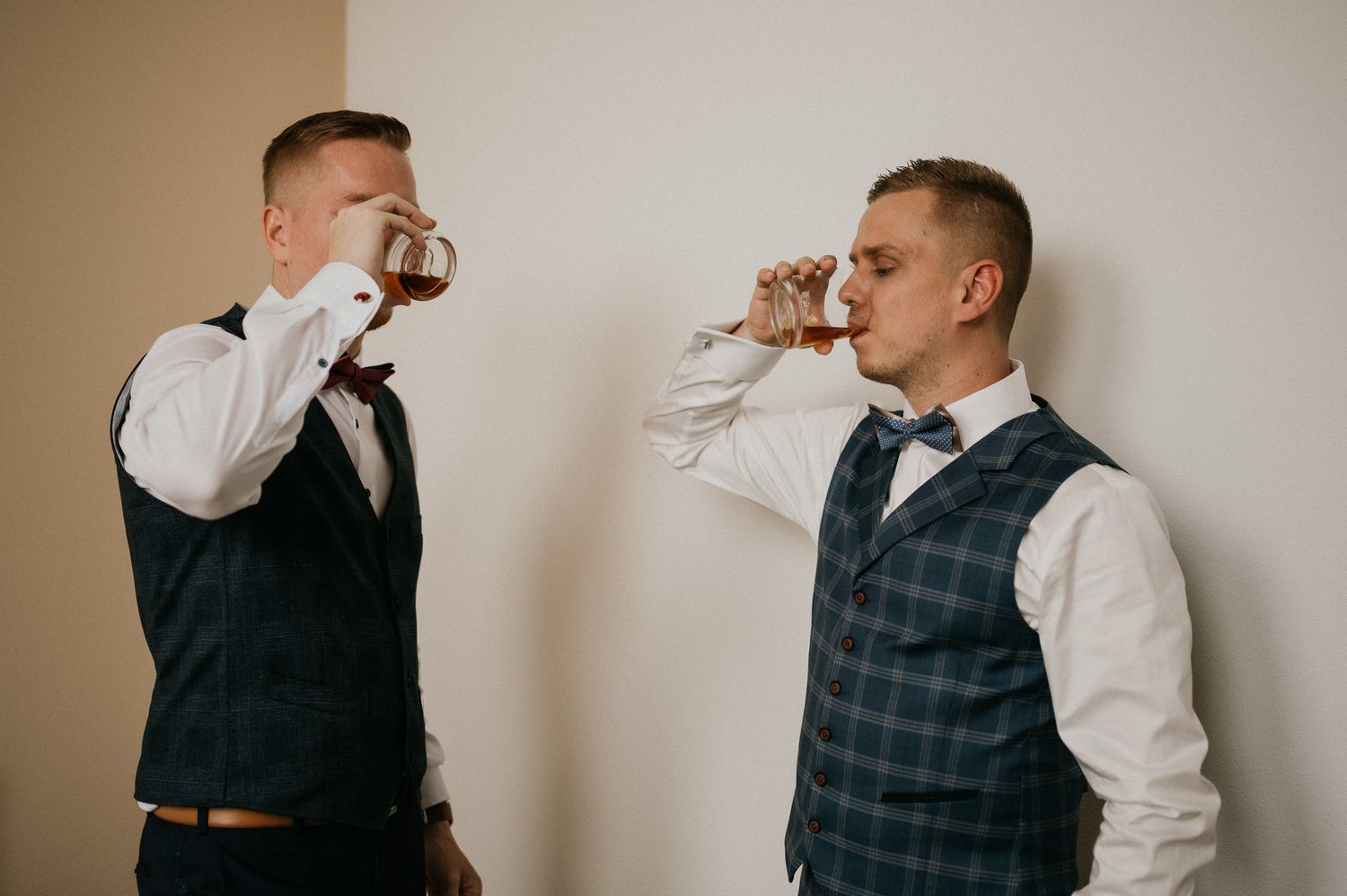 Two groomsmen in vests and dress shirts taking celebratory drinks together.