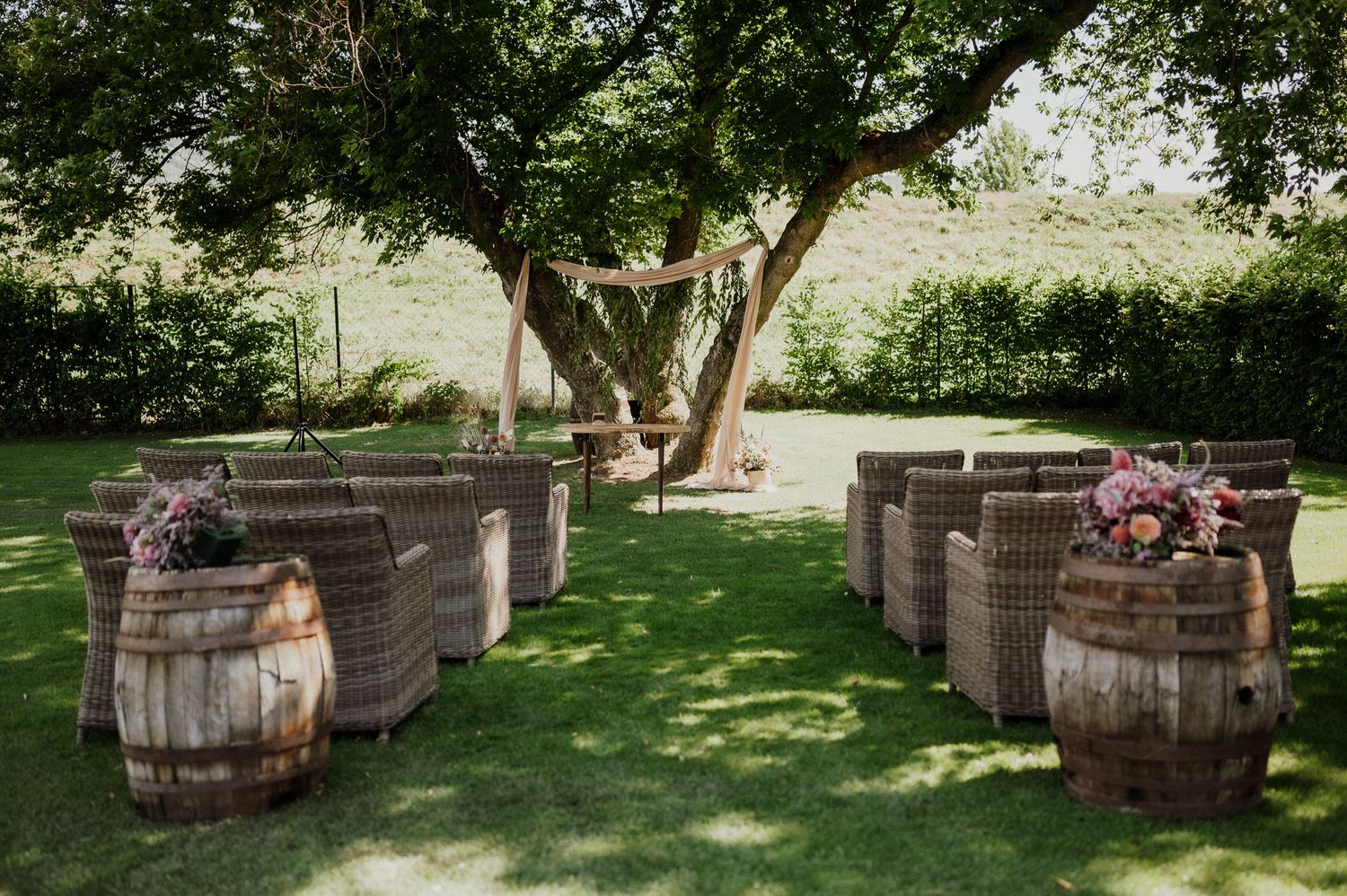 Garden wedding venue setup with rustic wine barrels and wicker chairs under large tree.