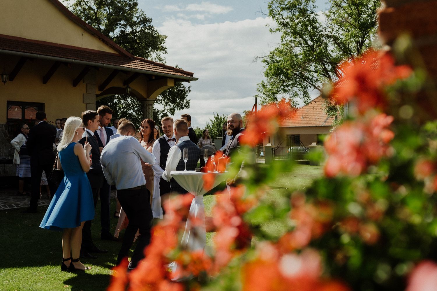 Wedding guests gather outside venue with vibrant orange flowers in foreground.