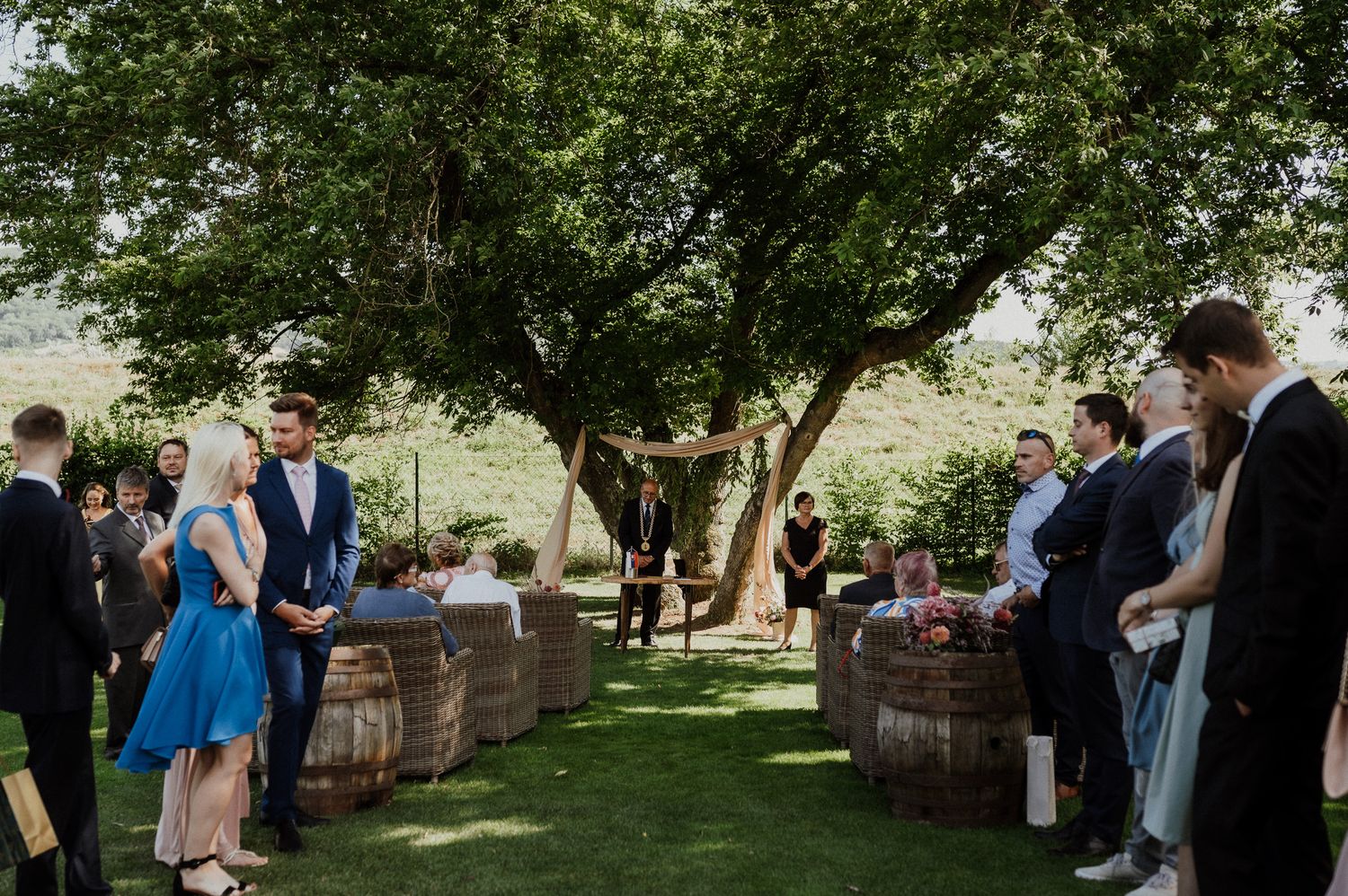 Wedding guests mingle in garden setting with rustic barrel decor and trees.