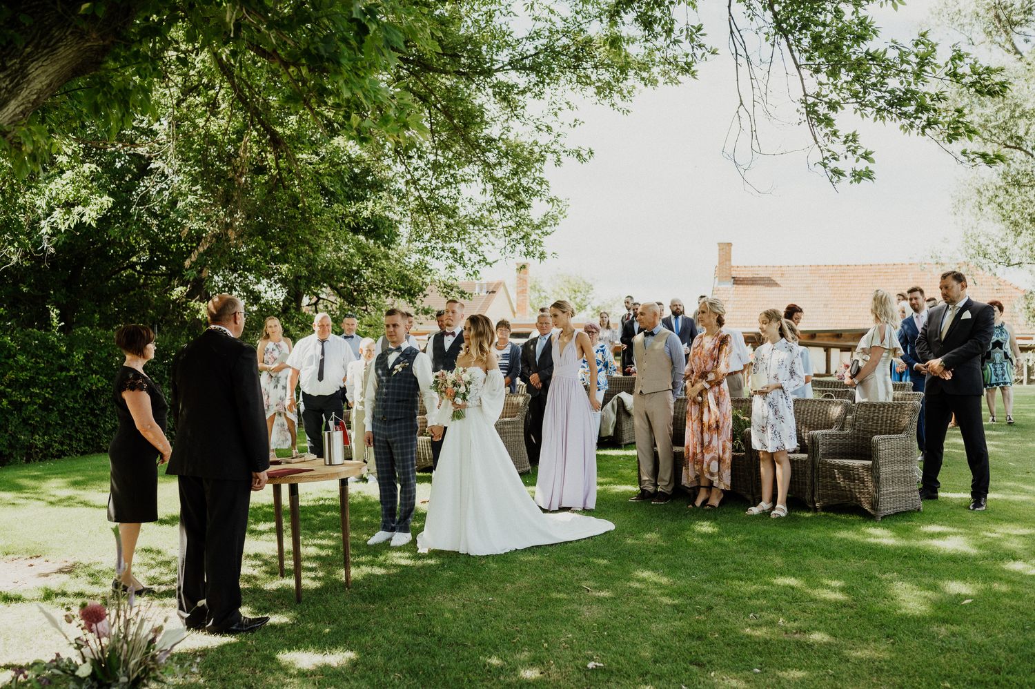Wedding ceremony guests gather on a sunlit lawn under trees with elegant outdoor decor.