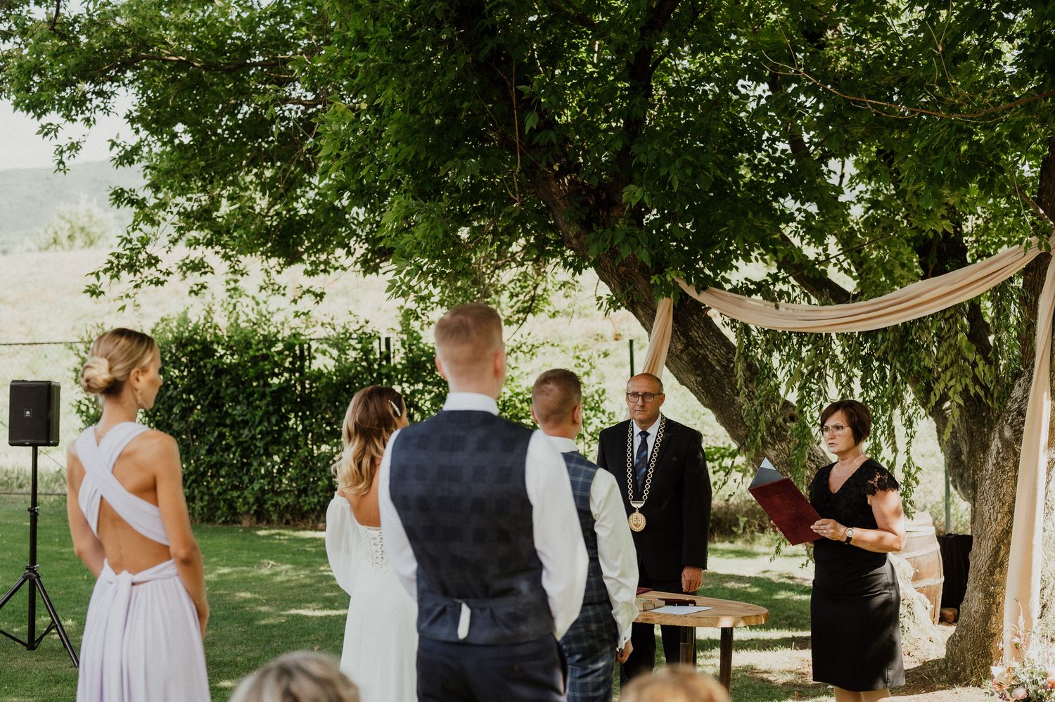 An outdoor wedding ceremony takes place under large trees with rustic decorations.