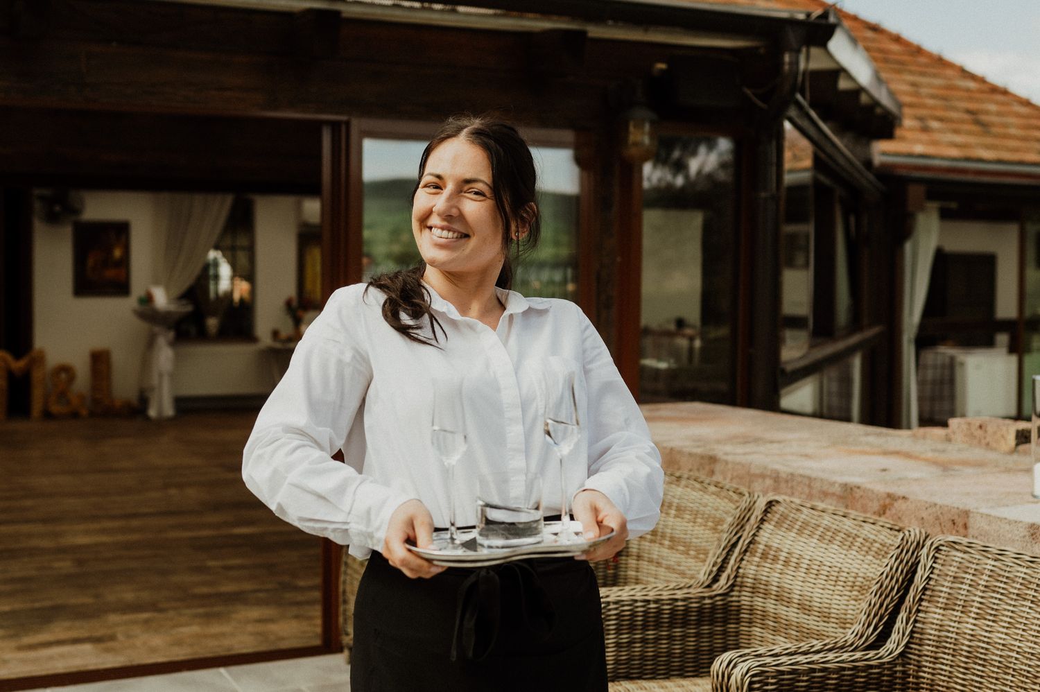 Server in white shirt carries drinks tray on outdoor wooden deck.