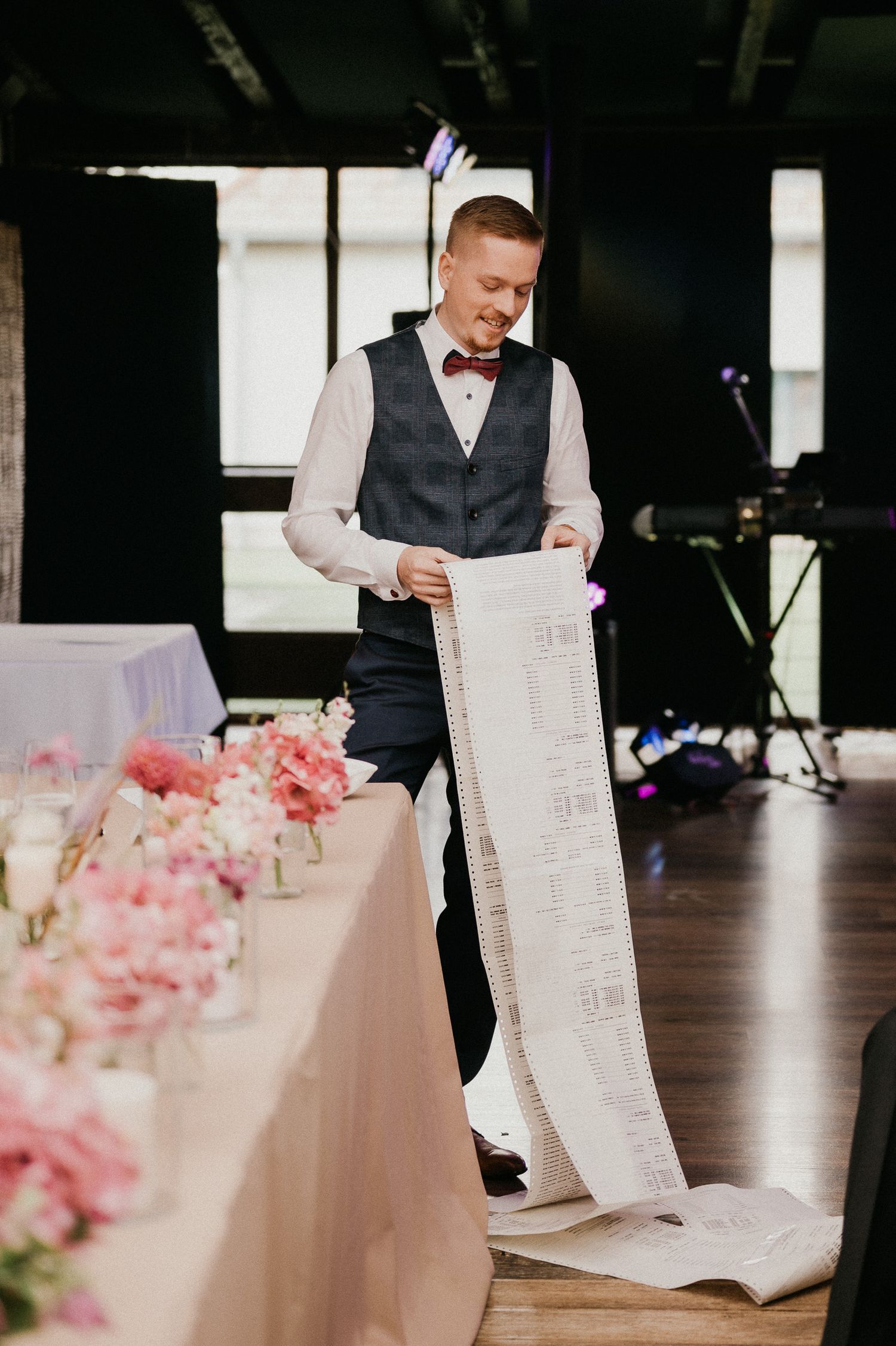 Wedding guest holding long scroll document at reception with pink floral centerpieces.