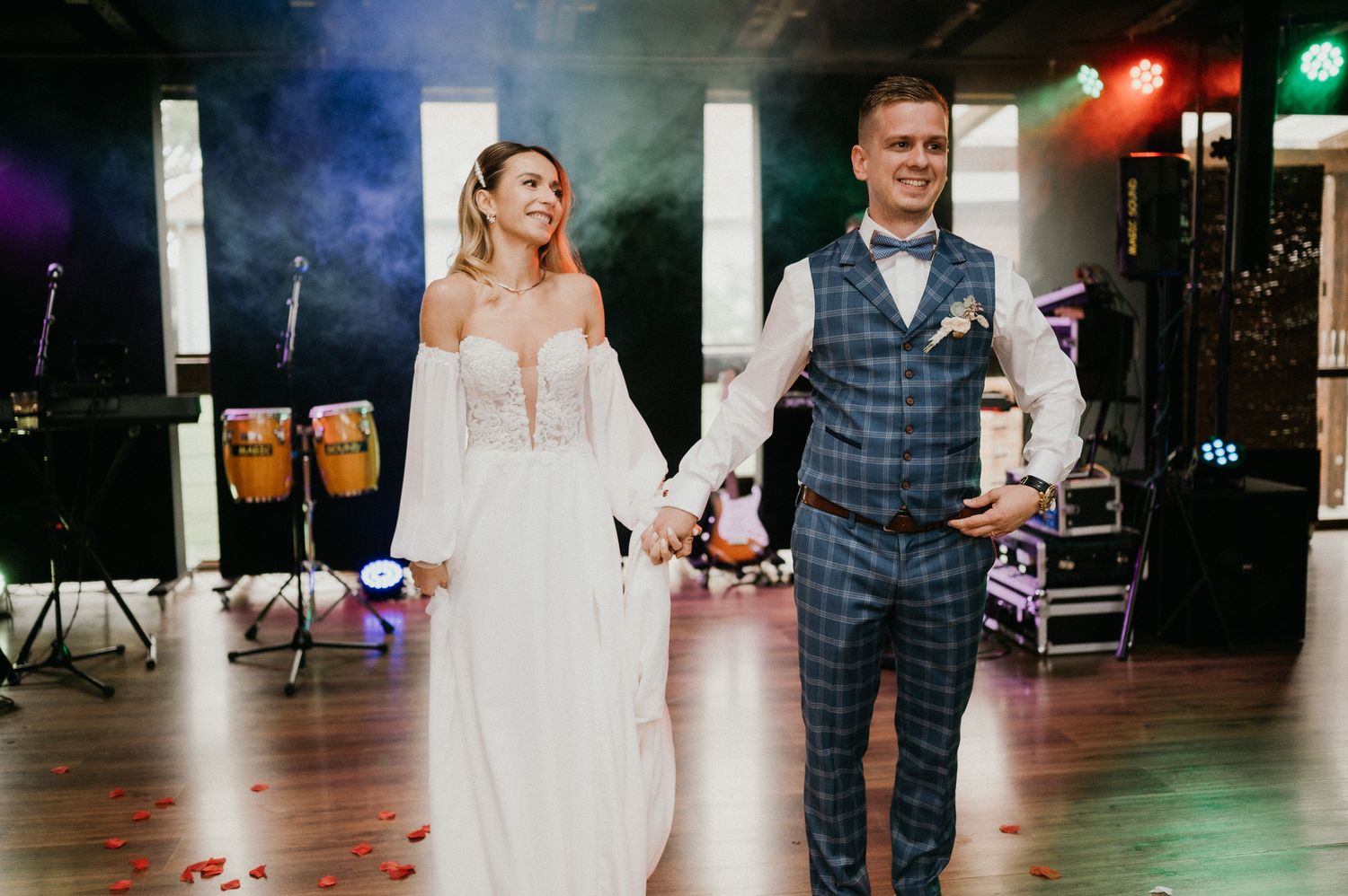 Newlyweds walking hand in hand at reception venue with stage lighting and smoke effects.
