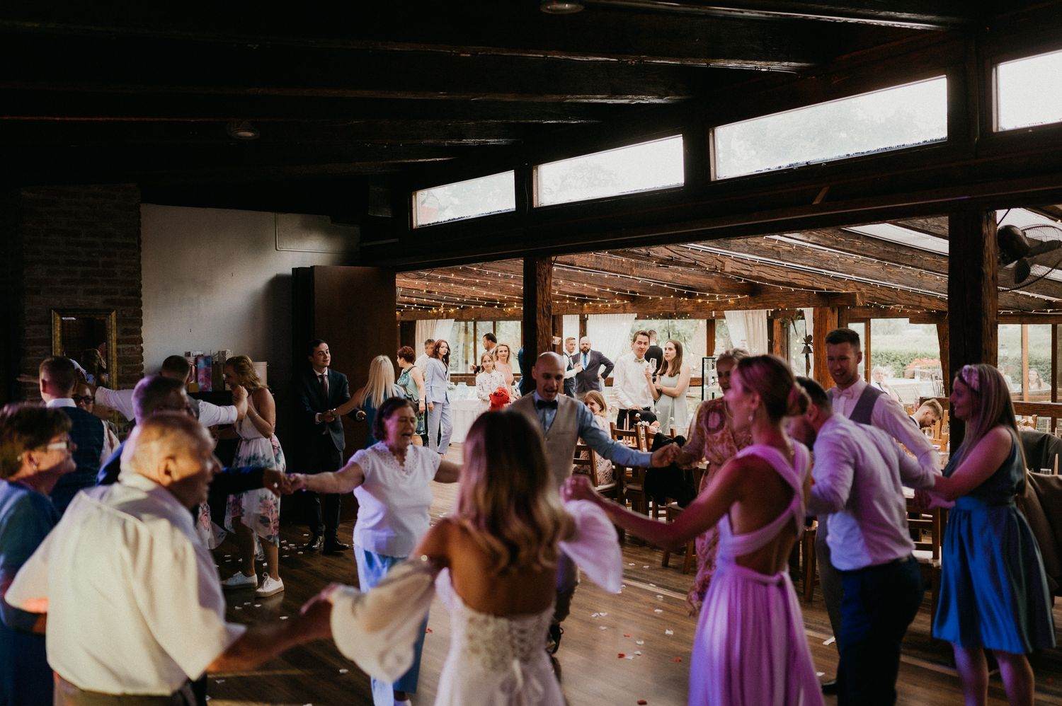 Wedding guests dancing at indoor reception with natural light streaming through windows.