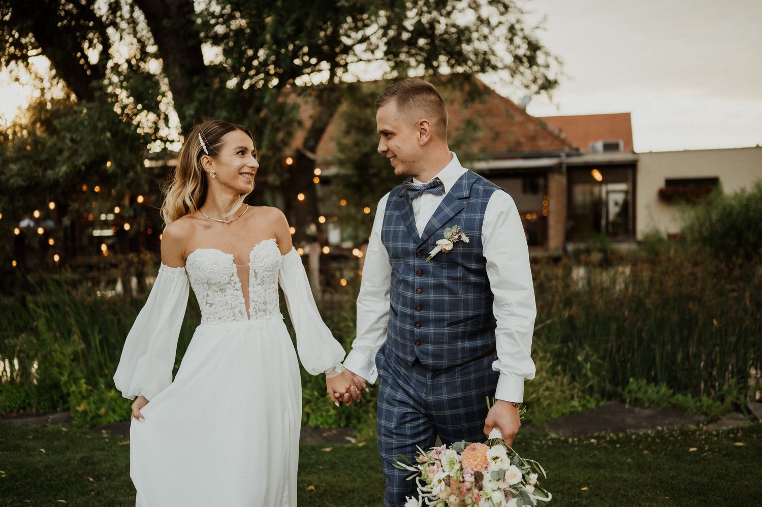 Couple walking together outdoors at sunset with rustic venue in background.