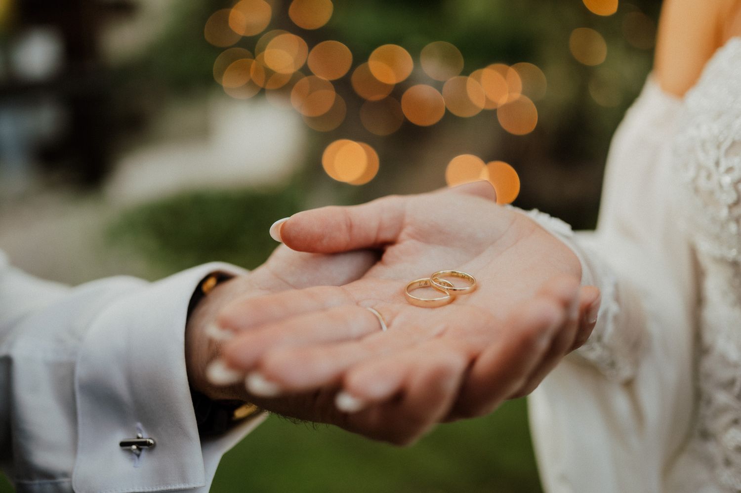 Close-up shot of wedding rings resting in open palms with bokeh lights in the background.