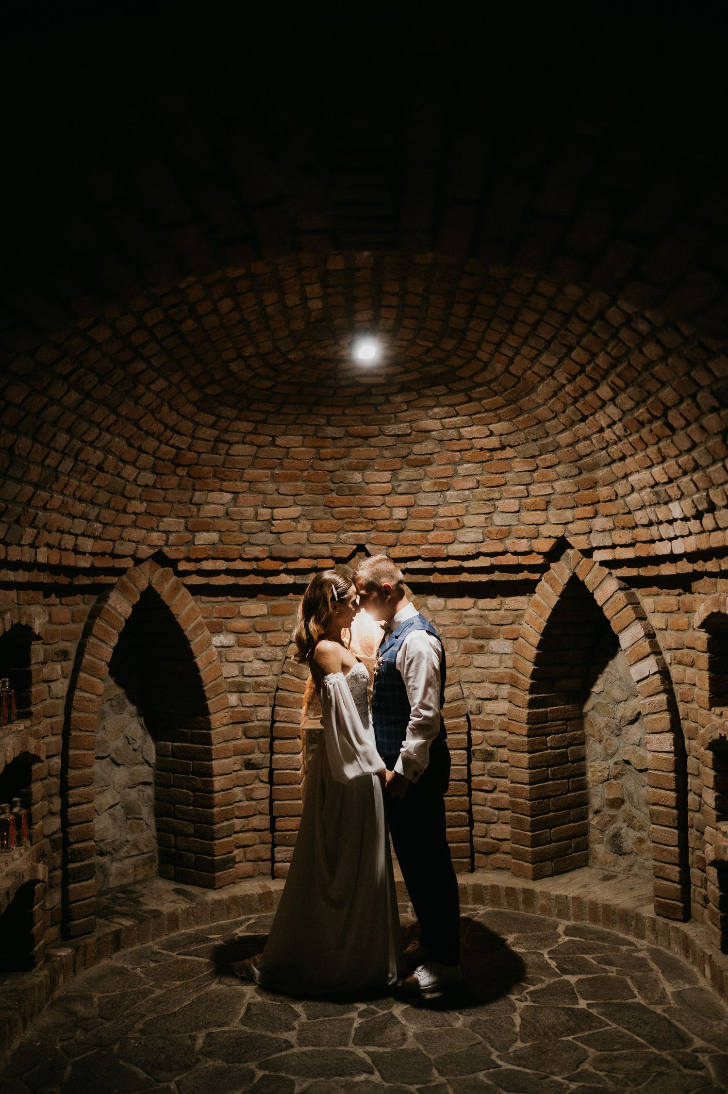 Romantic couple standing in a beautifully lit brick wine cellar with arched alcoves.