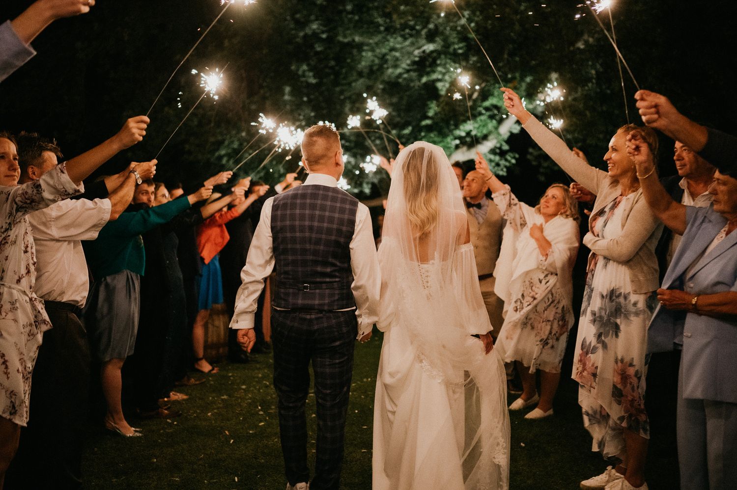 Wedding party holds sparklers forming illuminated archway during nighttime celebration.