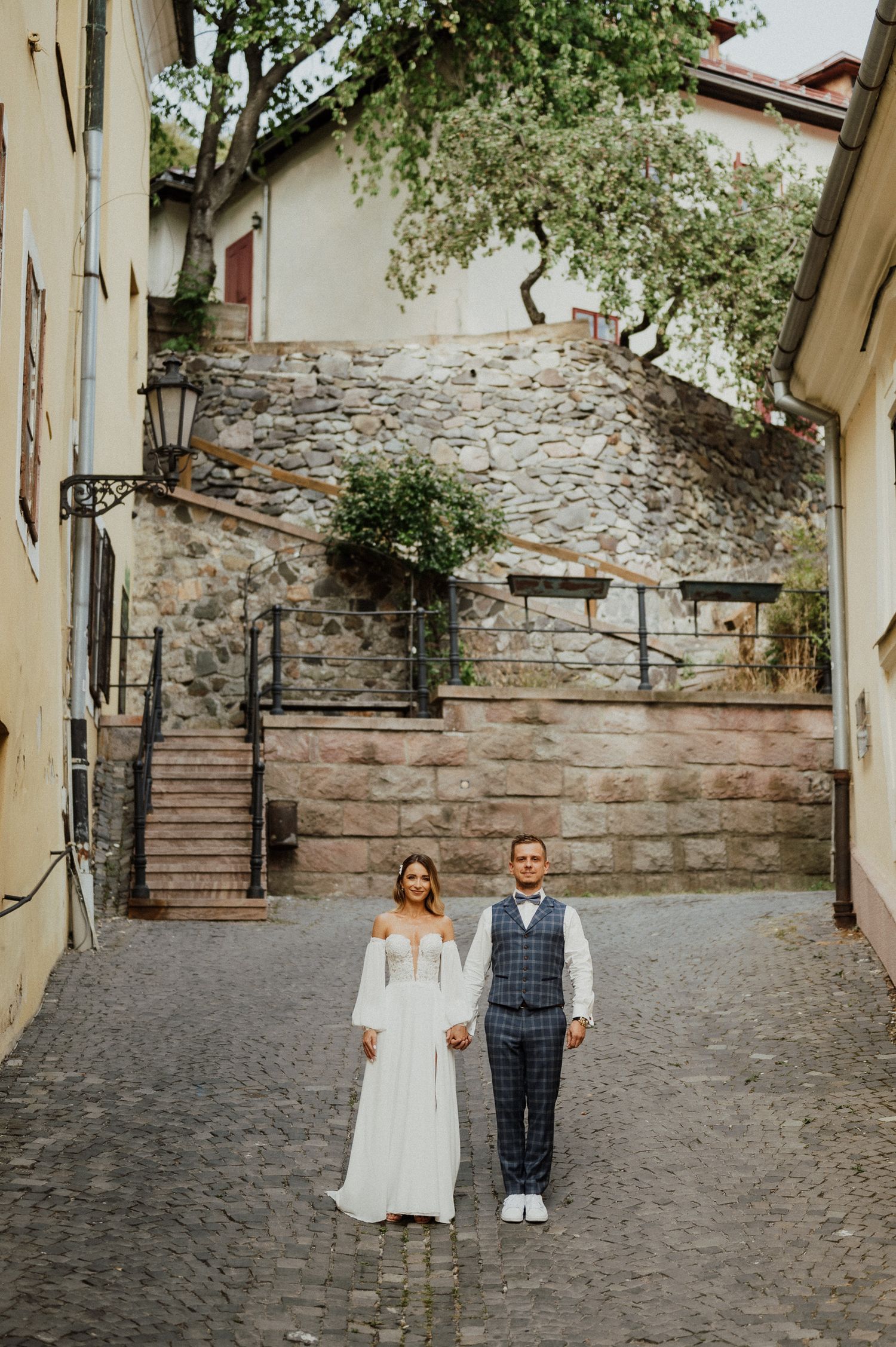 Couple stands on cobblestone street with historic stone stairs and walls in background.