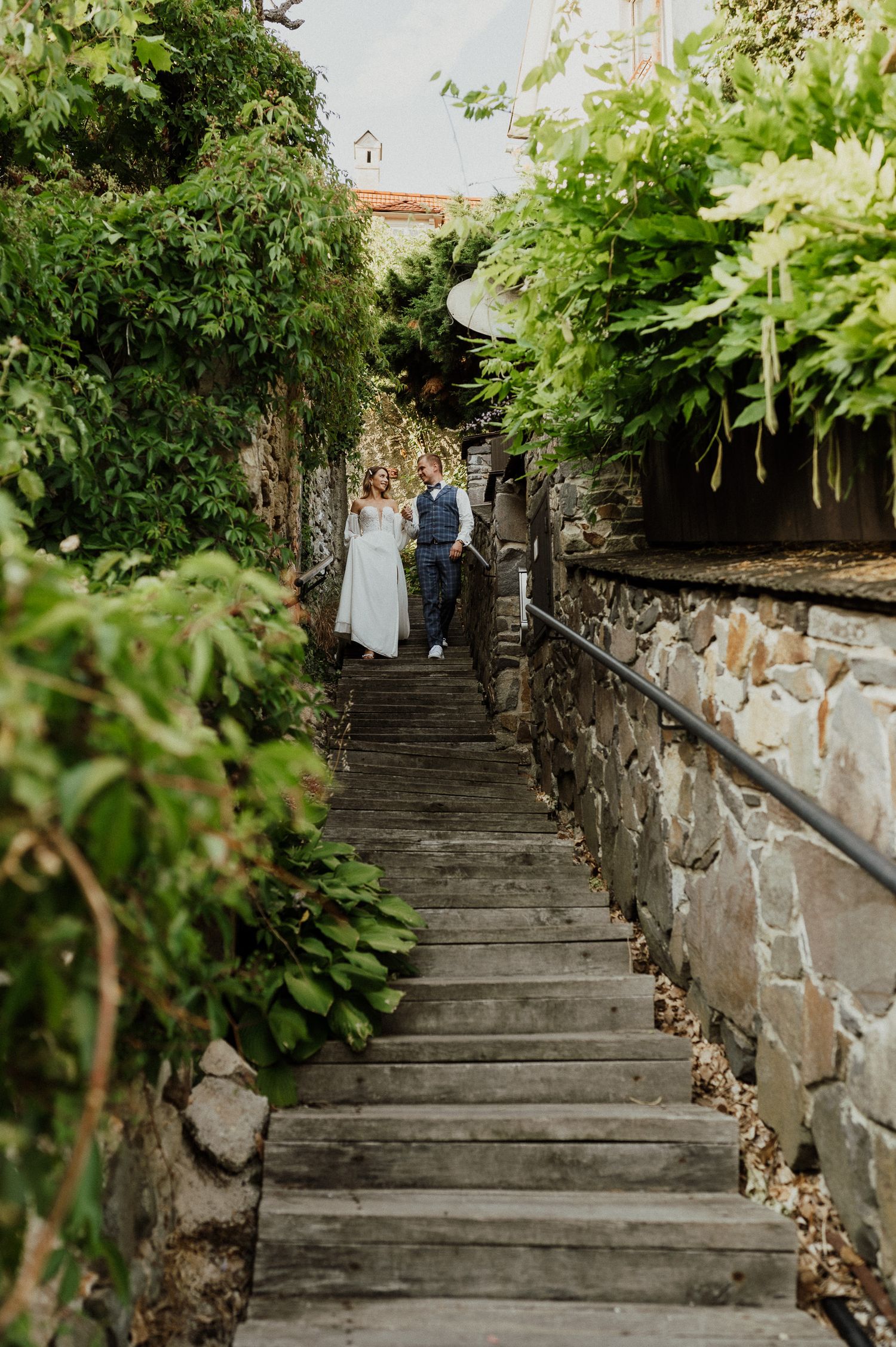 Couple descends stone steps surrounded by lush greenery and stone walls.