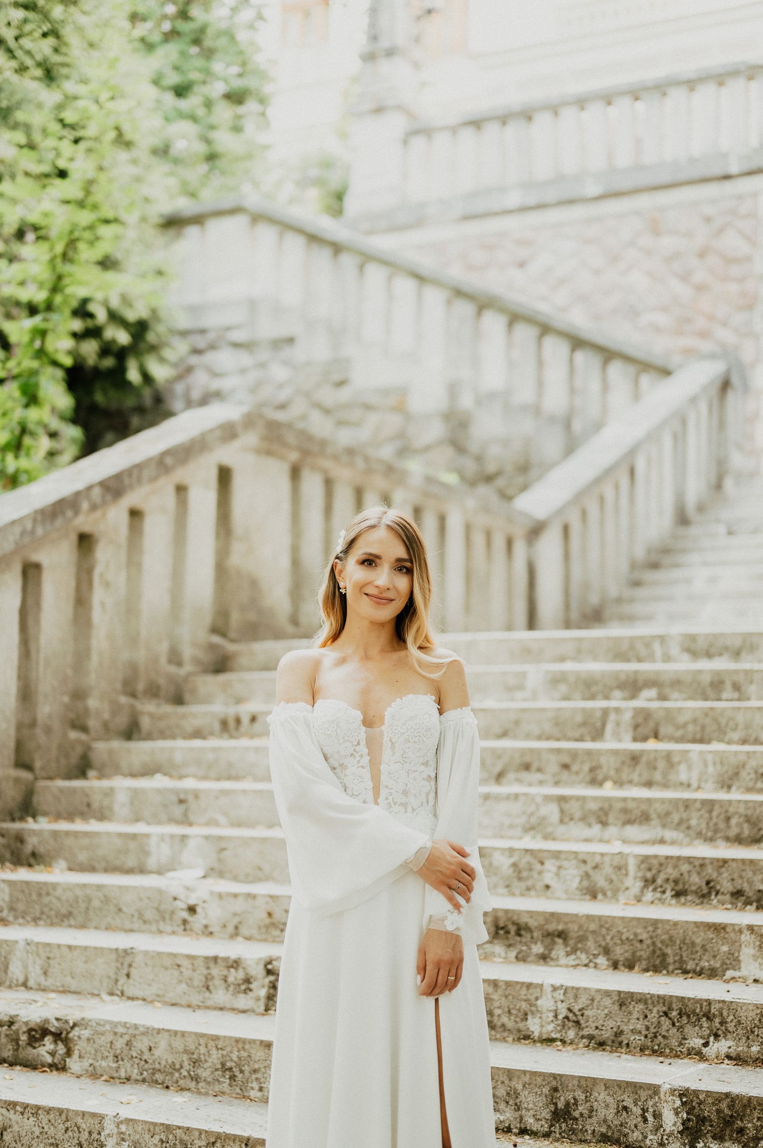 Elegant bride in off-shoulder white gown poses on grand stone staircase during outdoor wedding photoshoot.