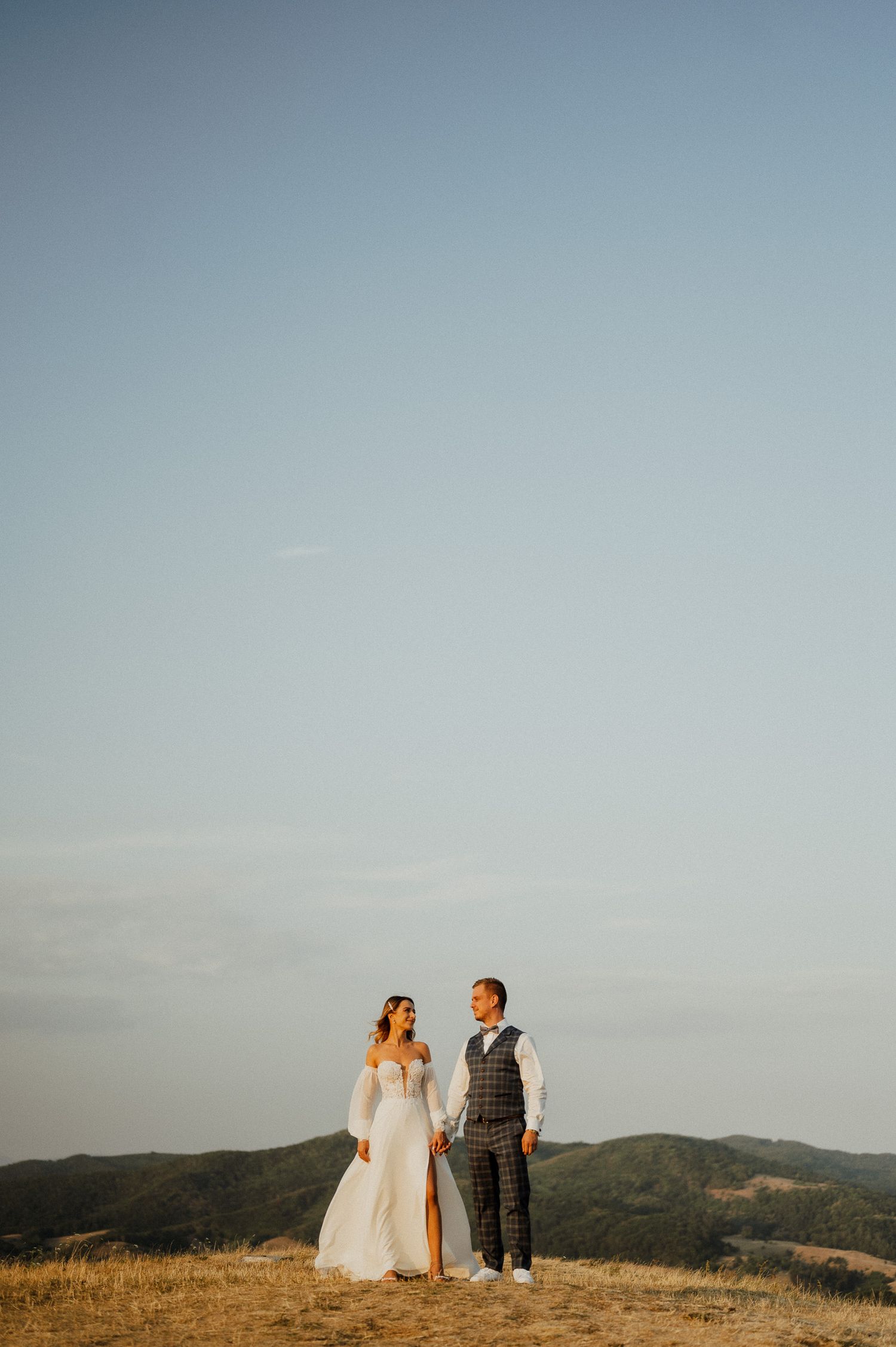 A couple stands together on a hilltop during golden hour in their wedding attire.