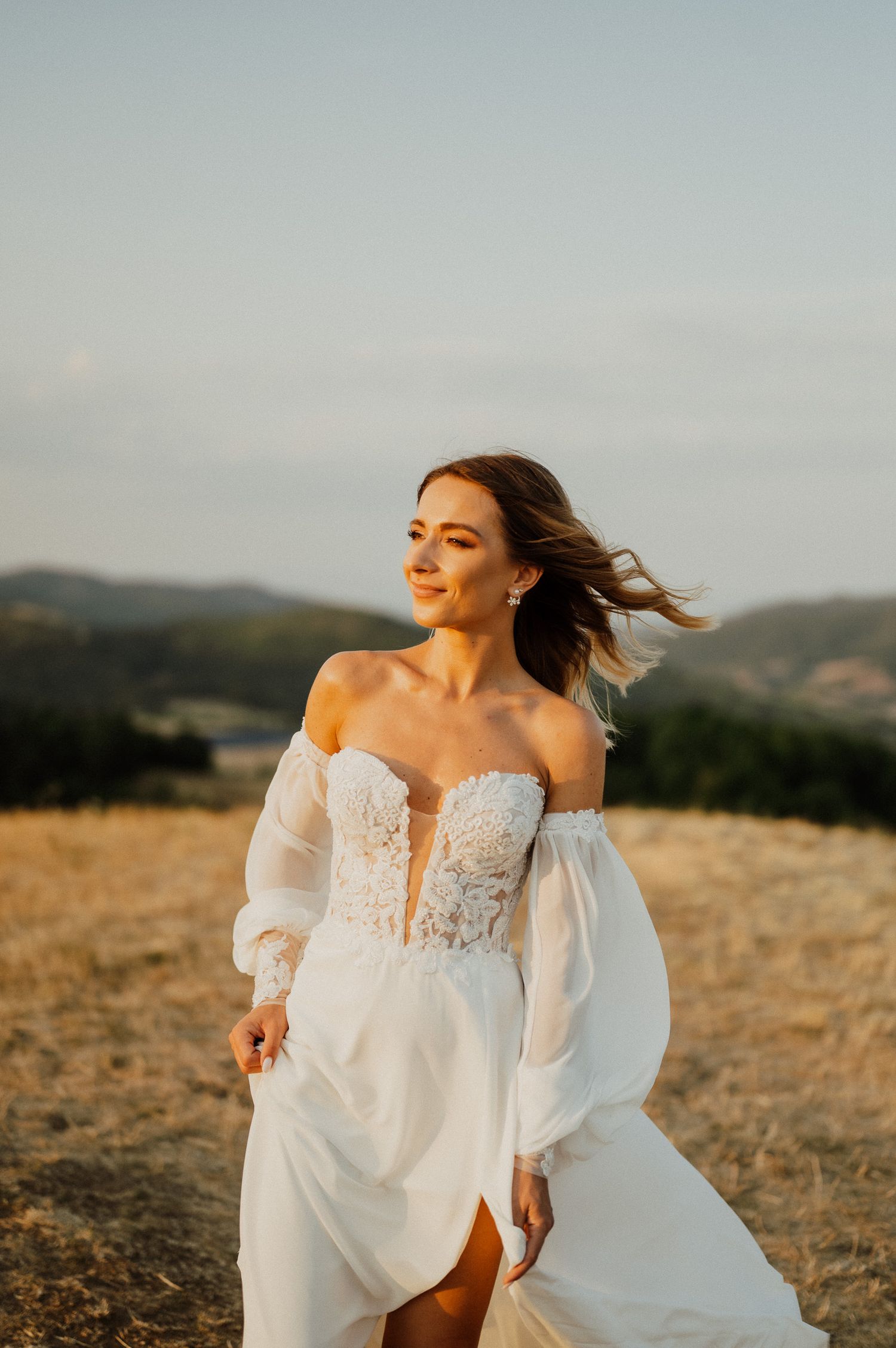 A bride in a flowing white dress poses in golden sunlight on a mountainside.