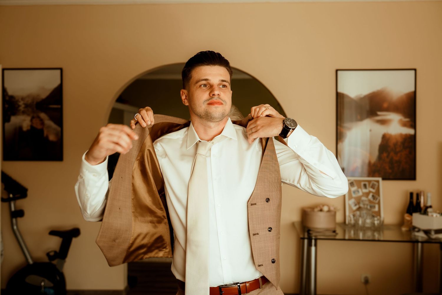 Person putting on a tan suit jacket while getting dressed in a room with framed artwork.
