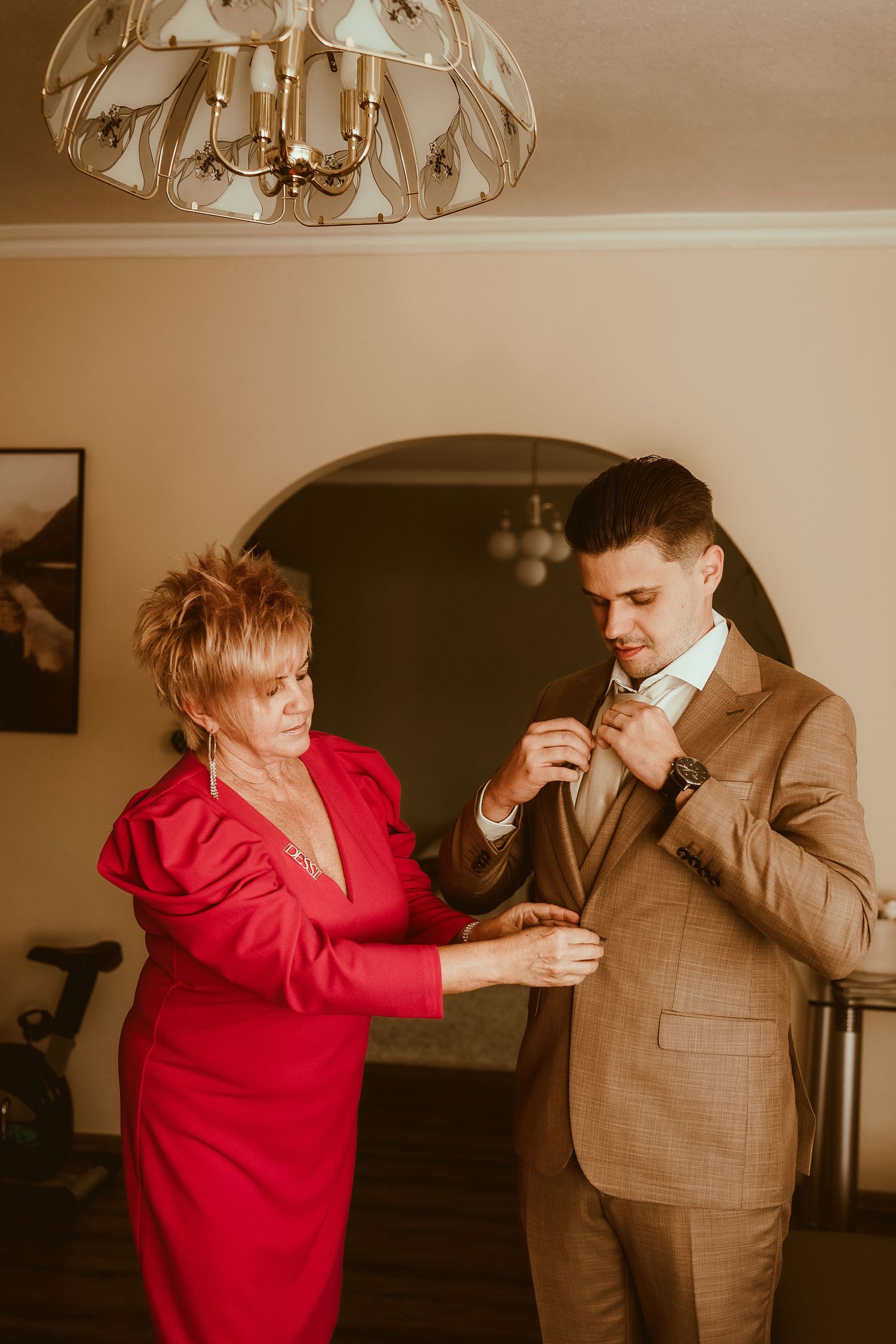 Woman in red helps adjust the tie of person wearing a tan suit during formal preparation.