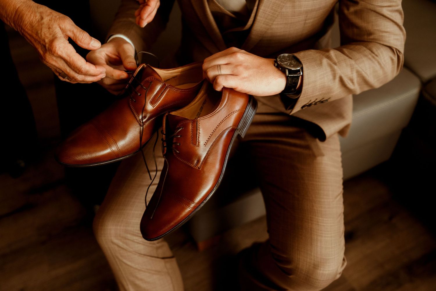 Close-up of hands examining brown leather dress shoes on someone wearing tan pants.