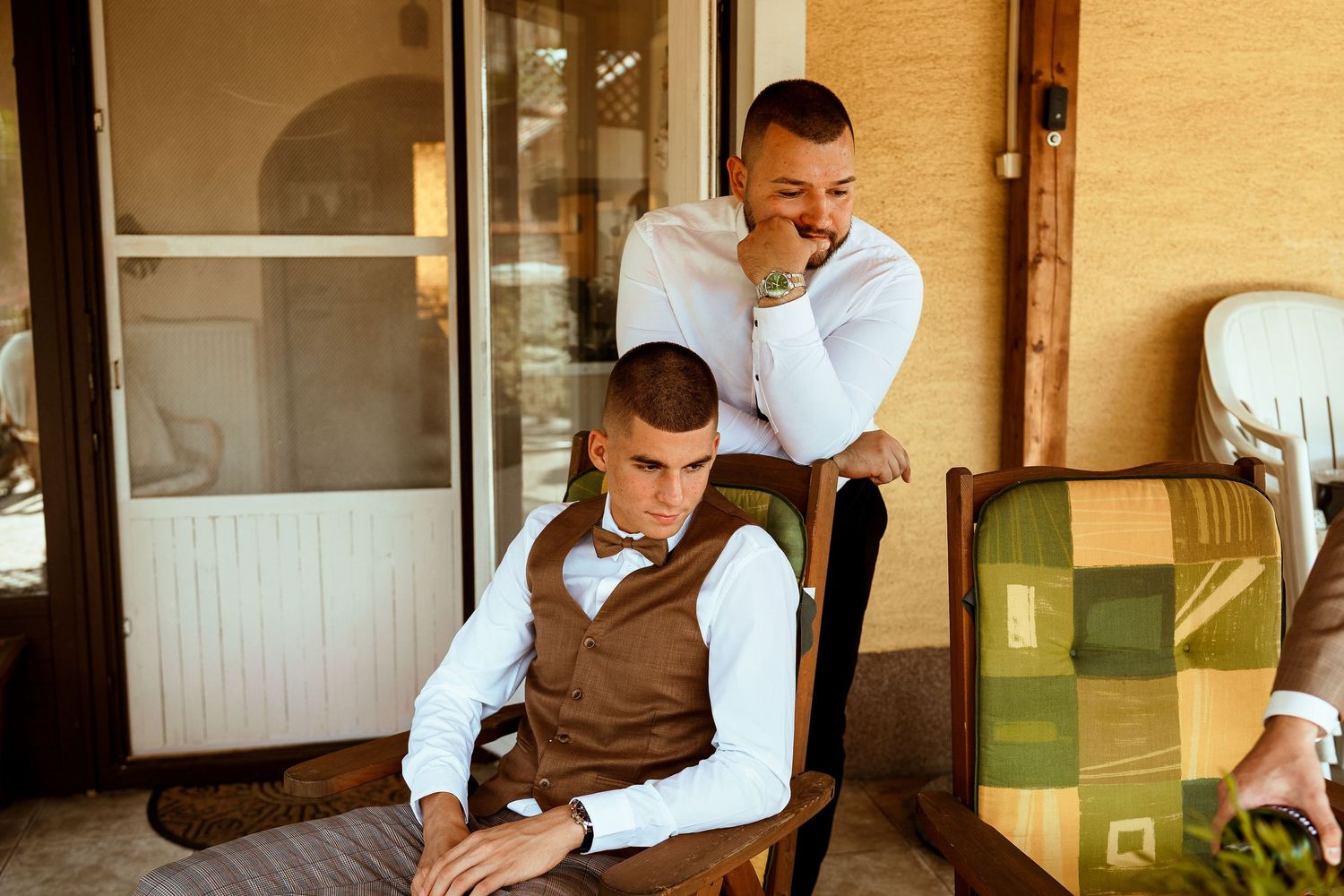 Two people in formal attire sitting and standing on a patio during a casual moment.
