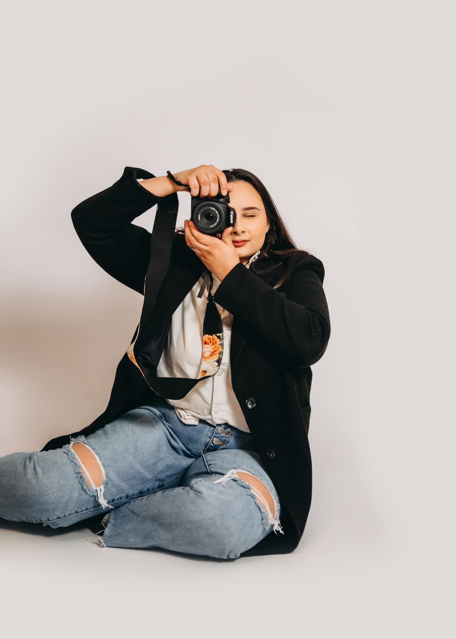 Photographer takes photo while sitting on floor wearing black coat and ripped jeans.
