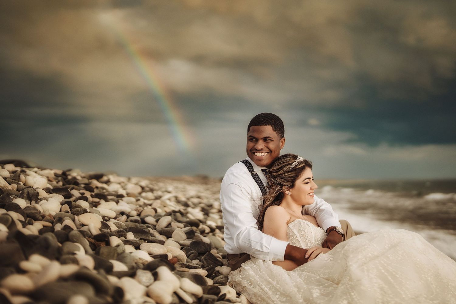 Wedding couple lounging on rocky beach with dramatic cloudy sky.