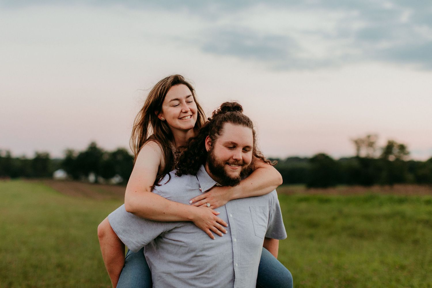 a guy carries his fiancee piggyback-style through a field at sunset