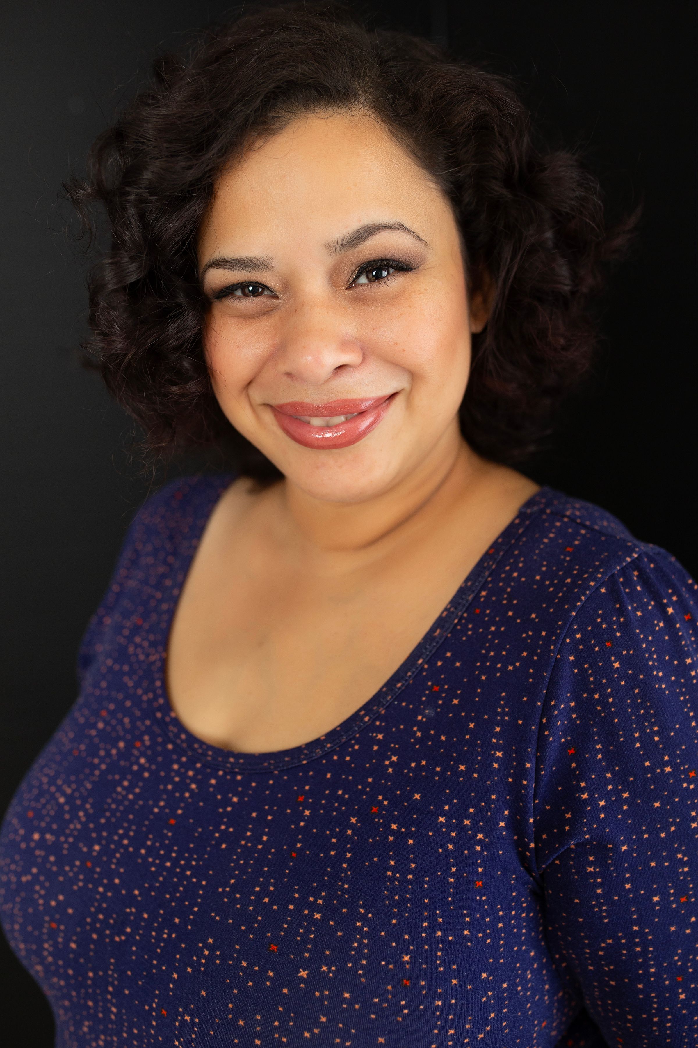 Professional headshot of person wearing navy sparkle dress against dark backdrop.