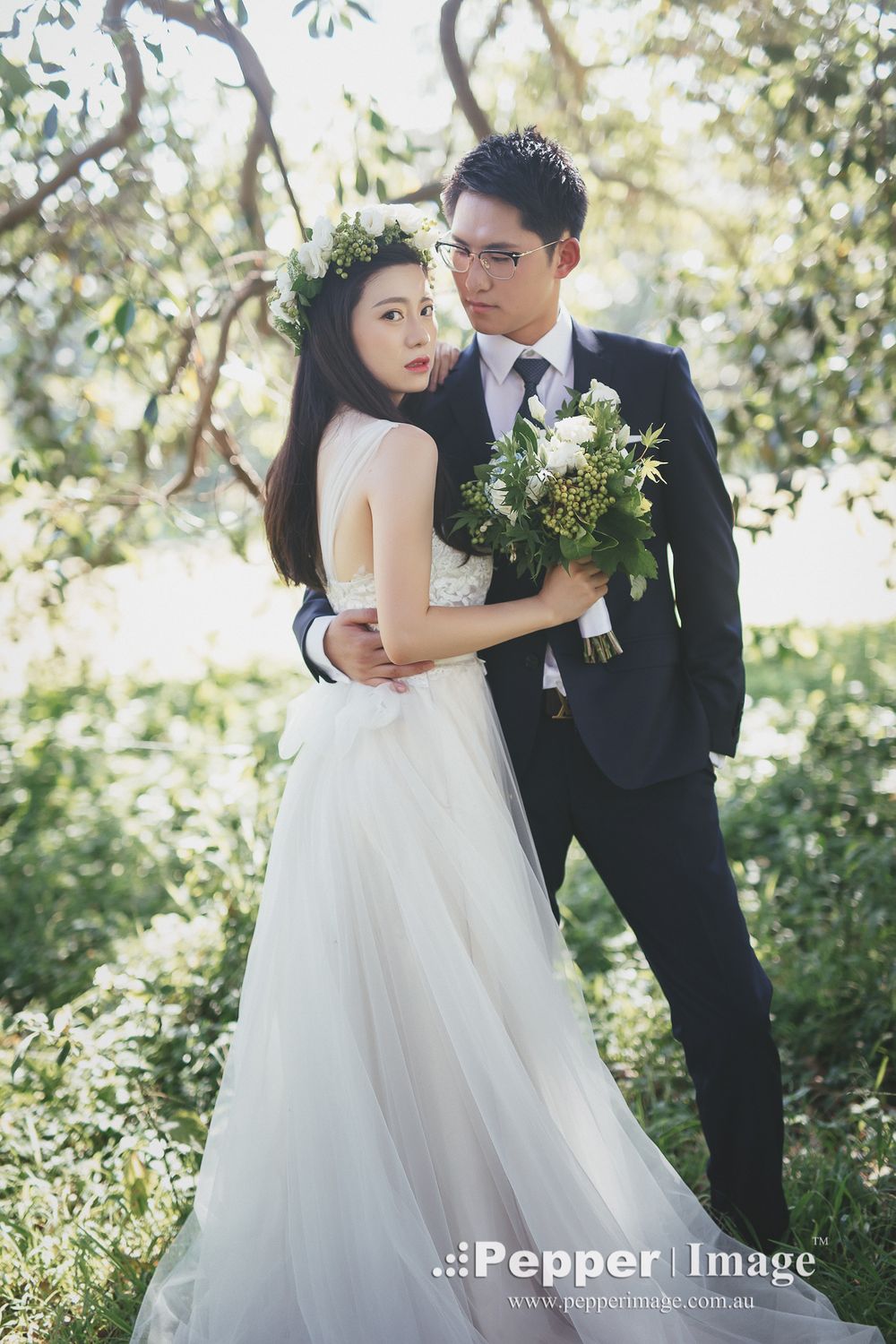 Bride wearing floral crown holds bouquet as groom embraces her in a sunlit outdoor garden setting.