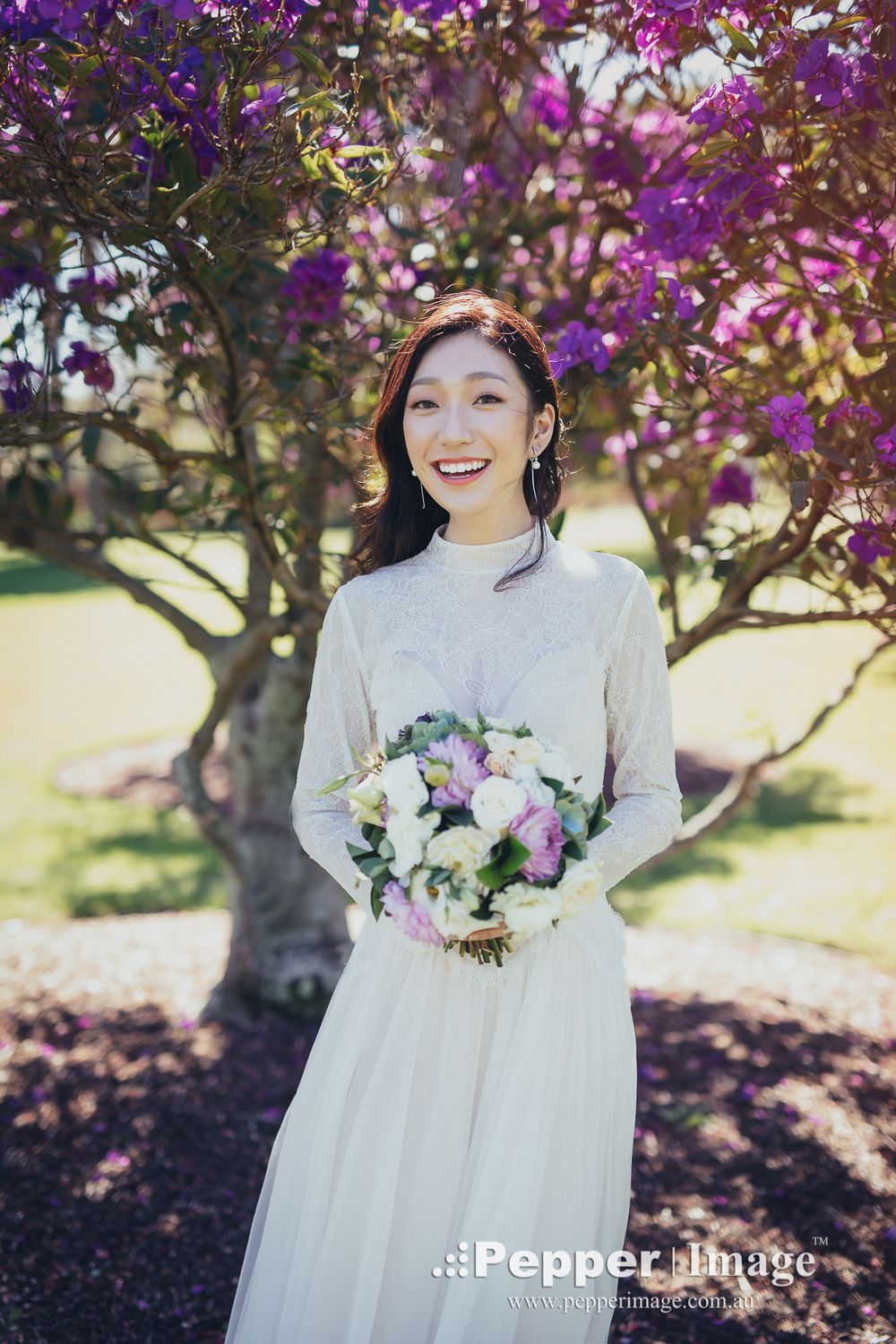 Smiling bride in ivory lace dress holding bouquet beside a vibrant purple flowering tree.