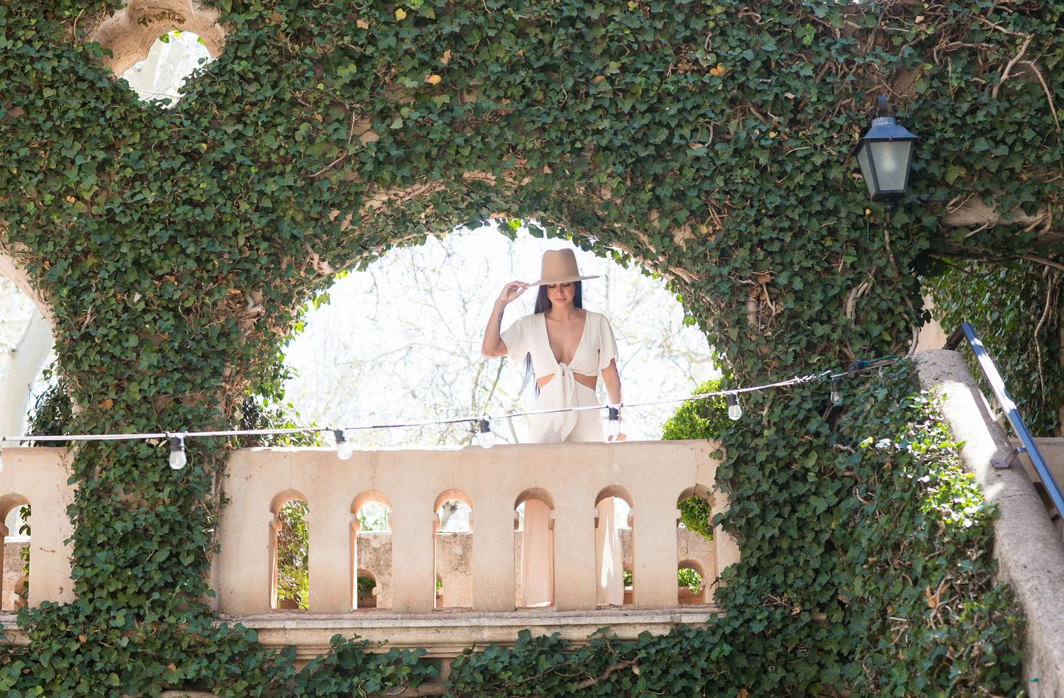 Elegant woman in white outfit and hat stands on ivy-covered balcony archway.