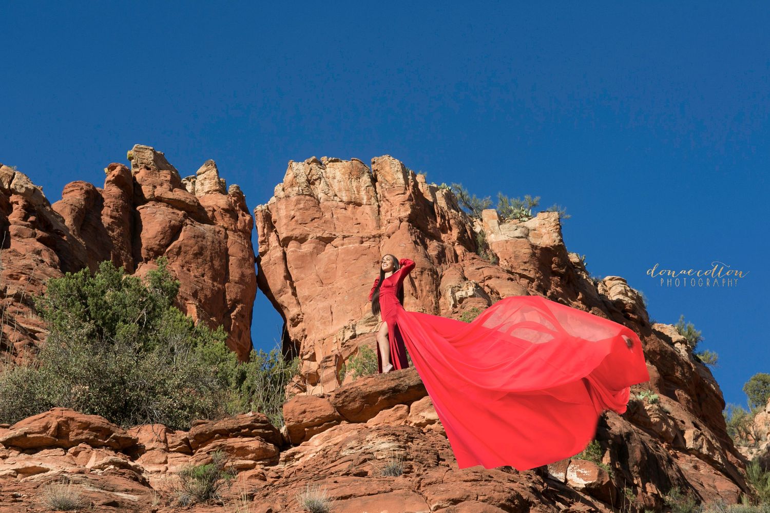 Woman in flowing red dress stands on red rock formations under vivid blue sky.