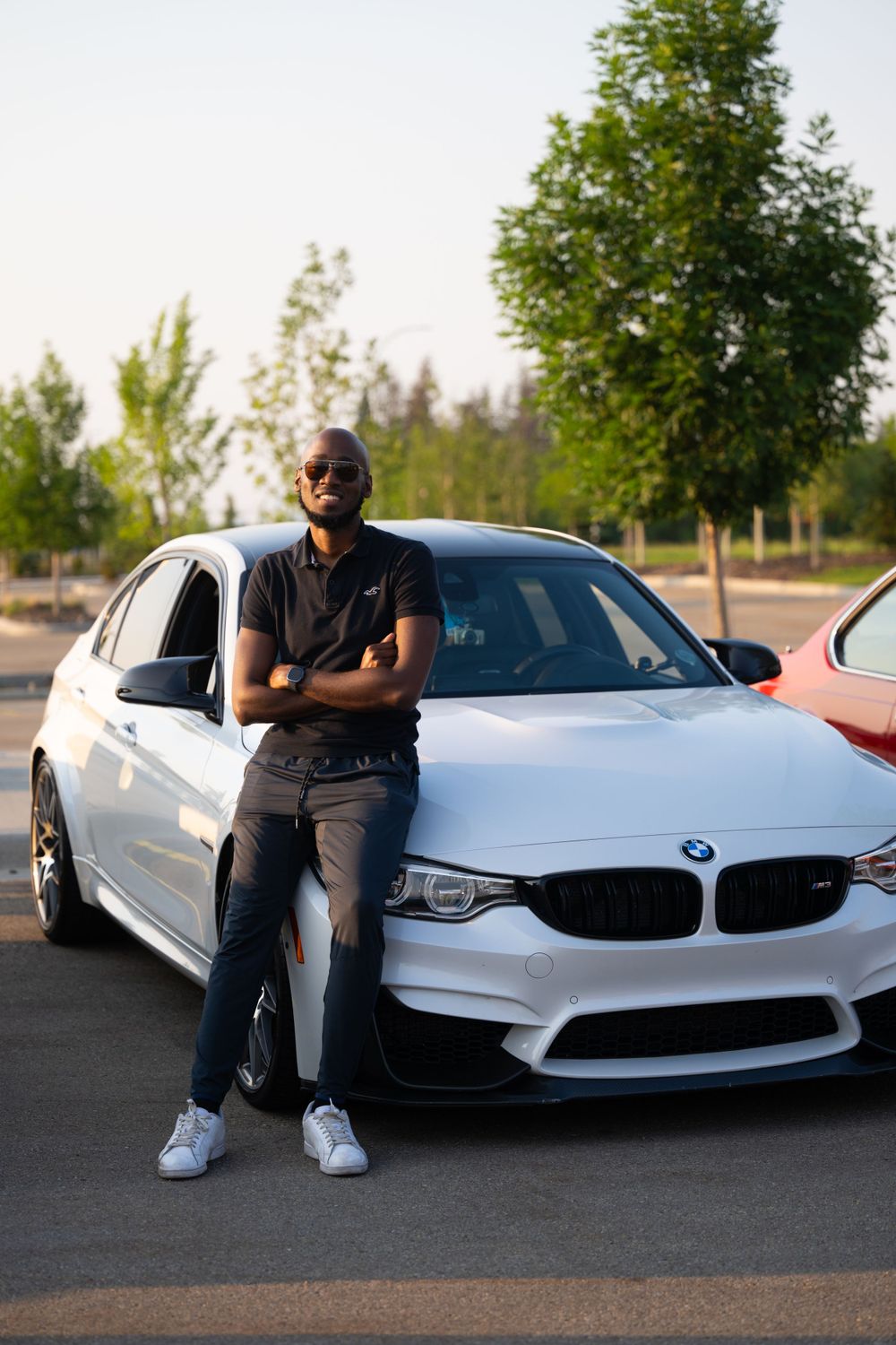 A white BMW M-series car is parked in a lot during sunset with someone sitting casually beside it.